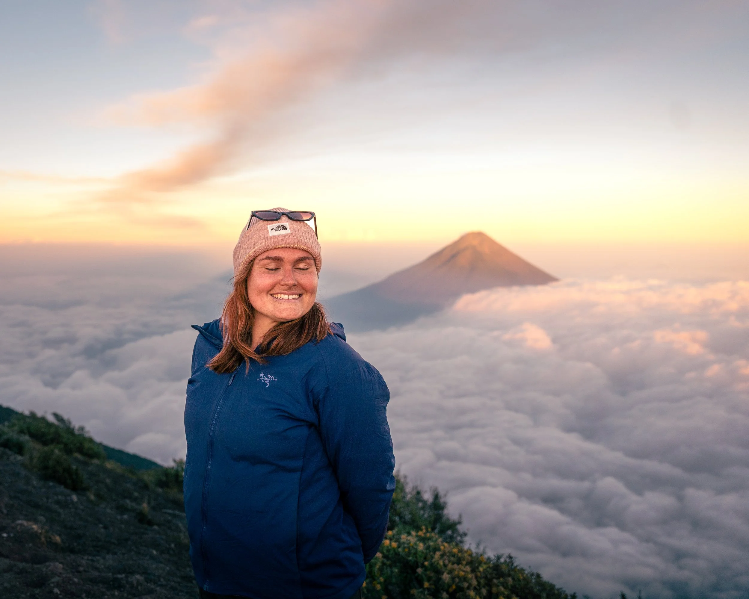 A woman with long hair wearing a pink beanie and sunglasses on her head smiles with eyes closed, standing on a mountain at sunrise or sunset with a mountain in the background and clouds below.