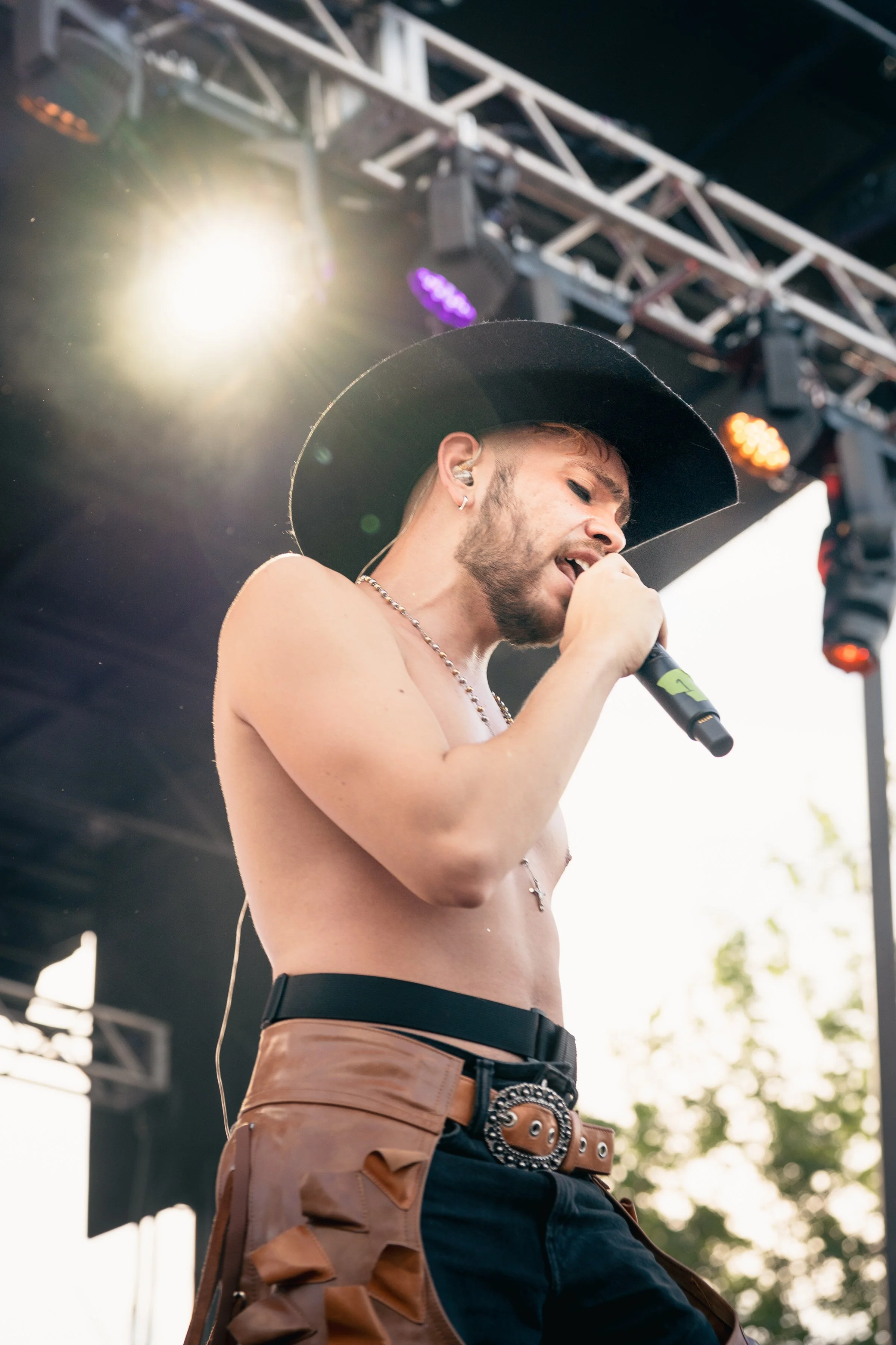 A shirtless male performer with a black wide-brim hat singing into a microphone on stage, with stage lights and metal scaffolding visible in the background.