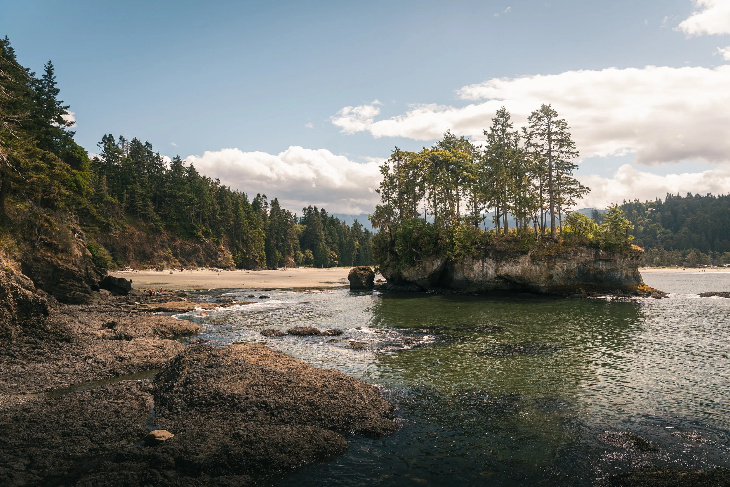 A coastal scene featuring rocky shoreline, green trees on small islands, and a partly cloudy sky.