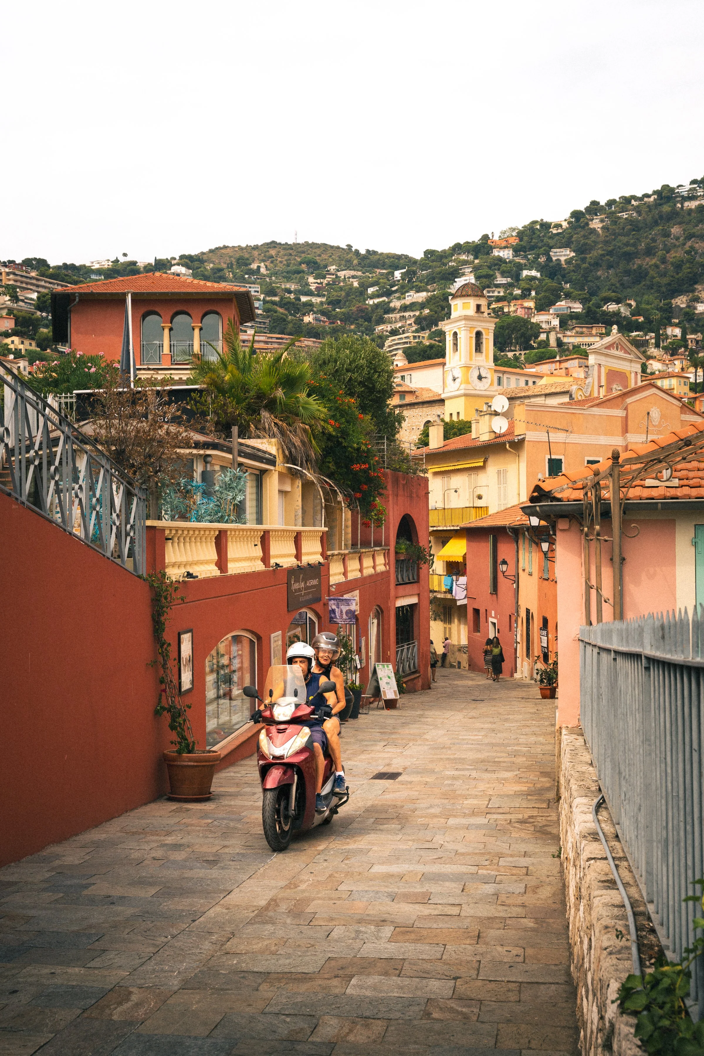 A street scene in a European village with colorful buildings, a clock tower, and a hilly backdrop. People are riding a scooter down the cobblestone street.