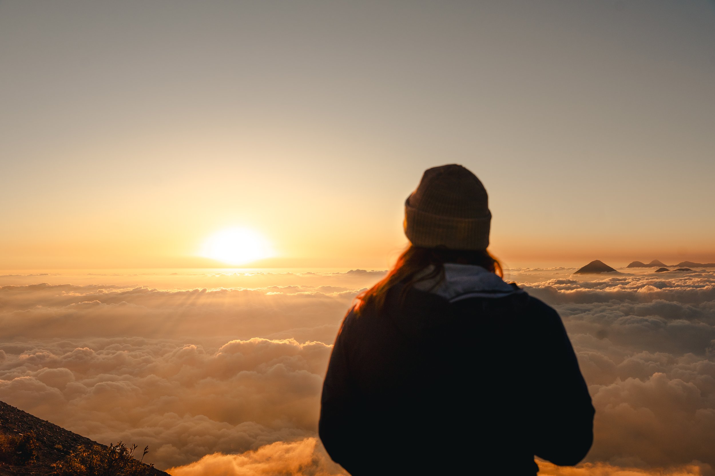 Person wearing a beanie looking at a sunrise or sunset above the clouds with mountains in the distance.