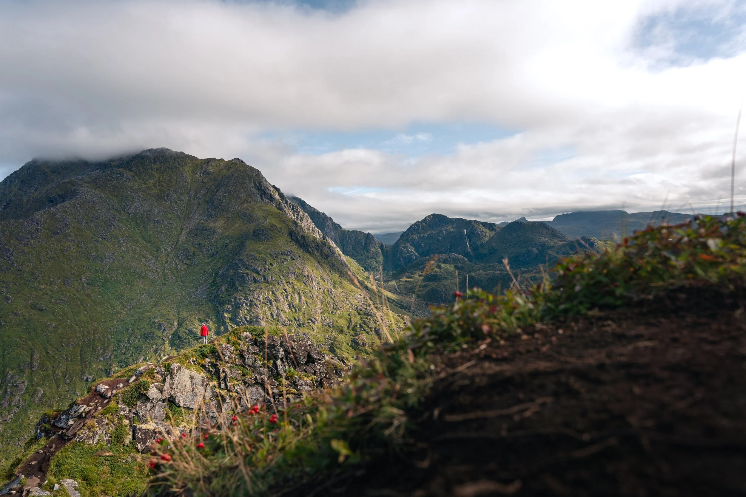 A person wearing a red jacket standing on a grassy ledge overlooking a mountainous landscape with clouds and blue sky above.