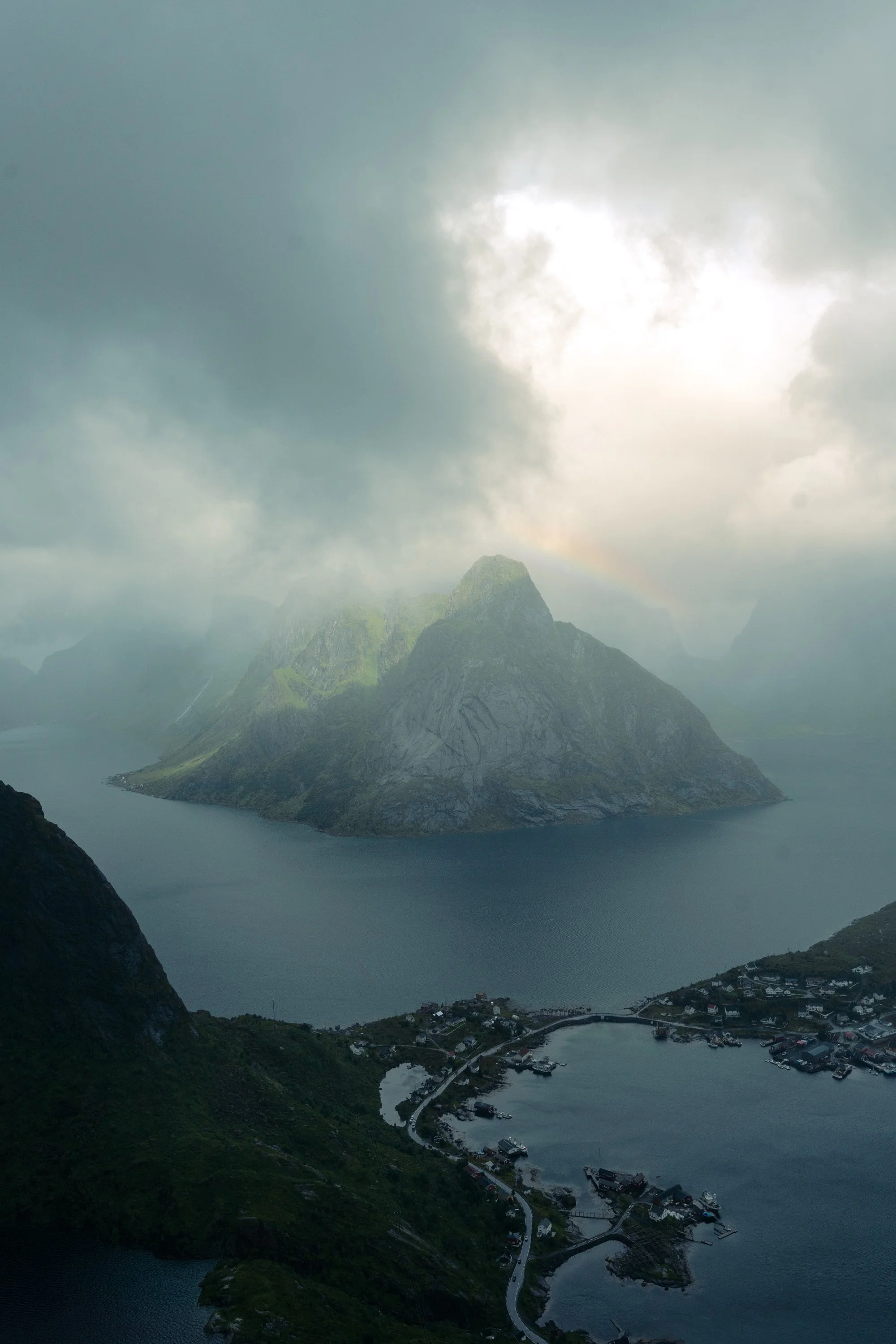 A scenic view of a fjord with a small village at the base of mountains, partially covered by low clouds and mist, with a rainbow faintly visible in the sky.