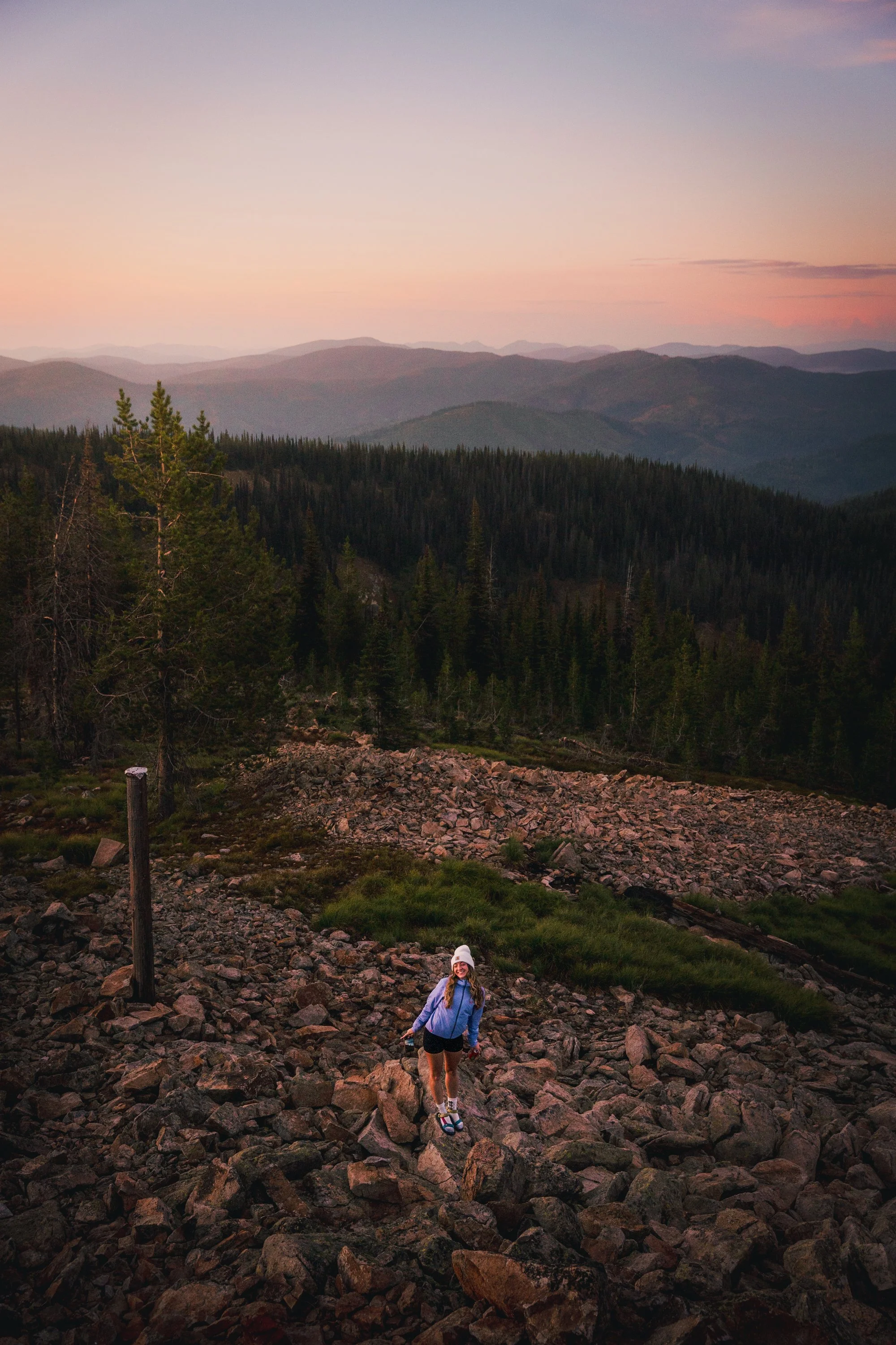 A woman in outdoor clothing, including a white hat and blue jacket, hiking on a rocky terrain with a scenic view of mountains and a colorful sky at sunset or dawn.
