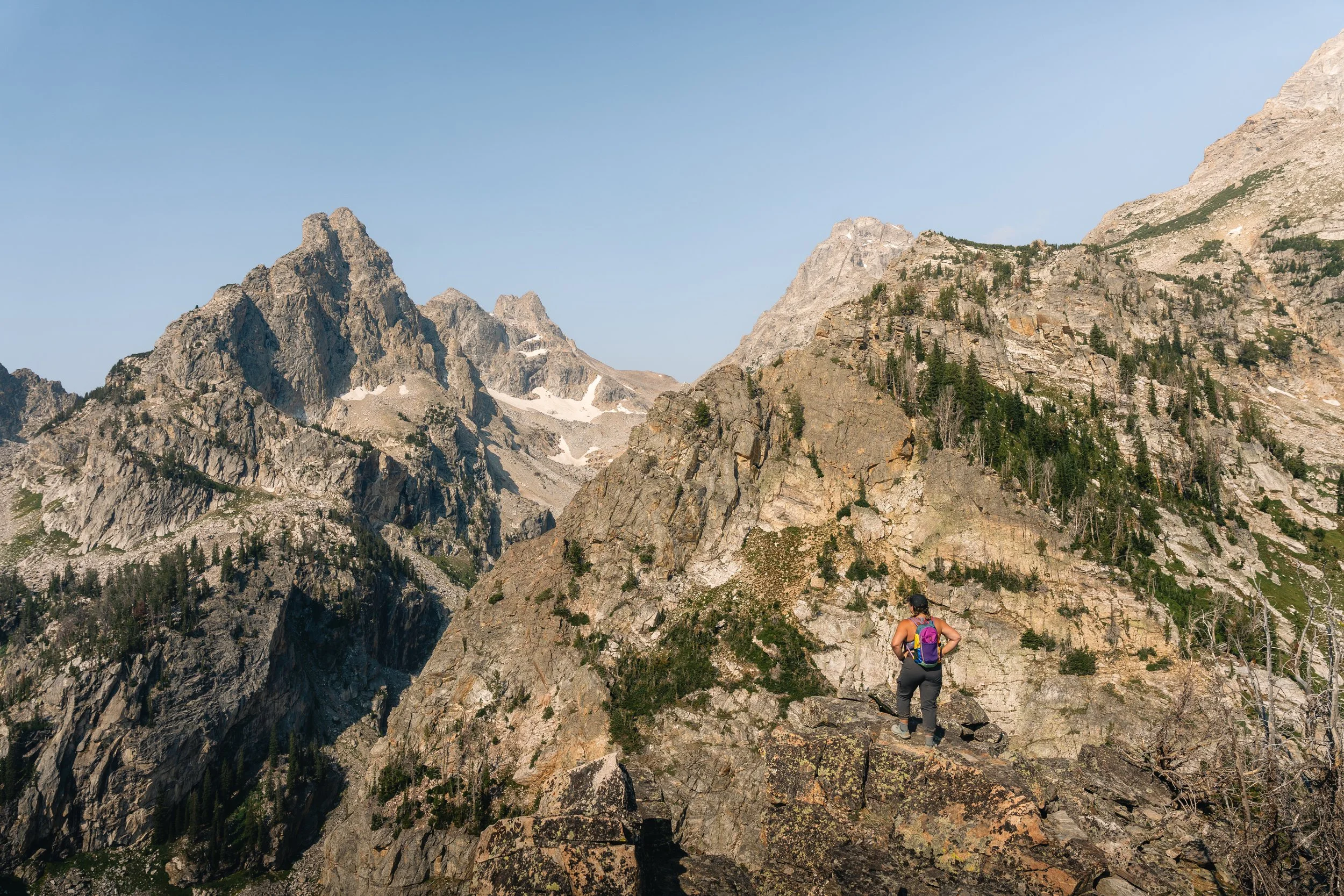 A person wearing a purple backpack hiking in a rocky mountain landscape with tall peaks and patches of snow under a clear blue sky.