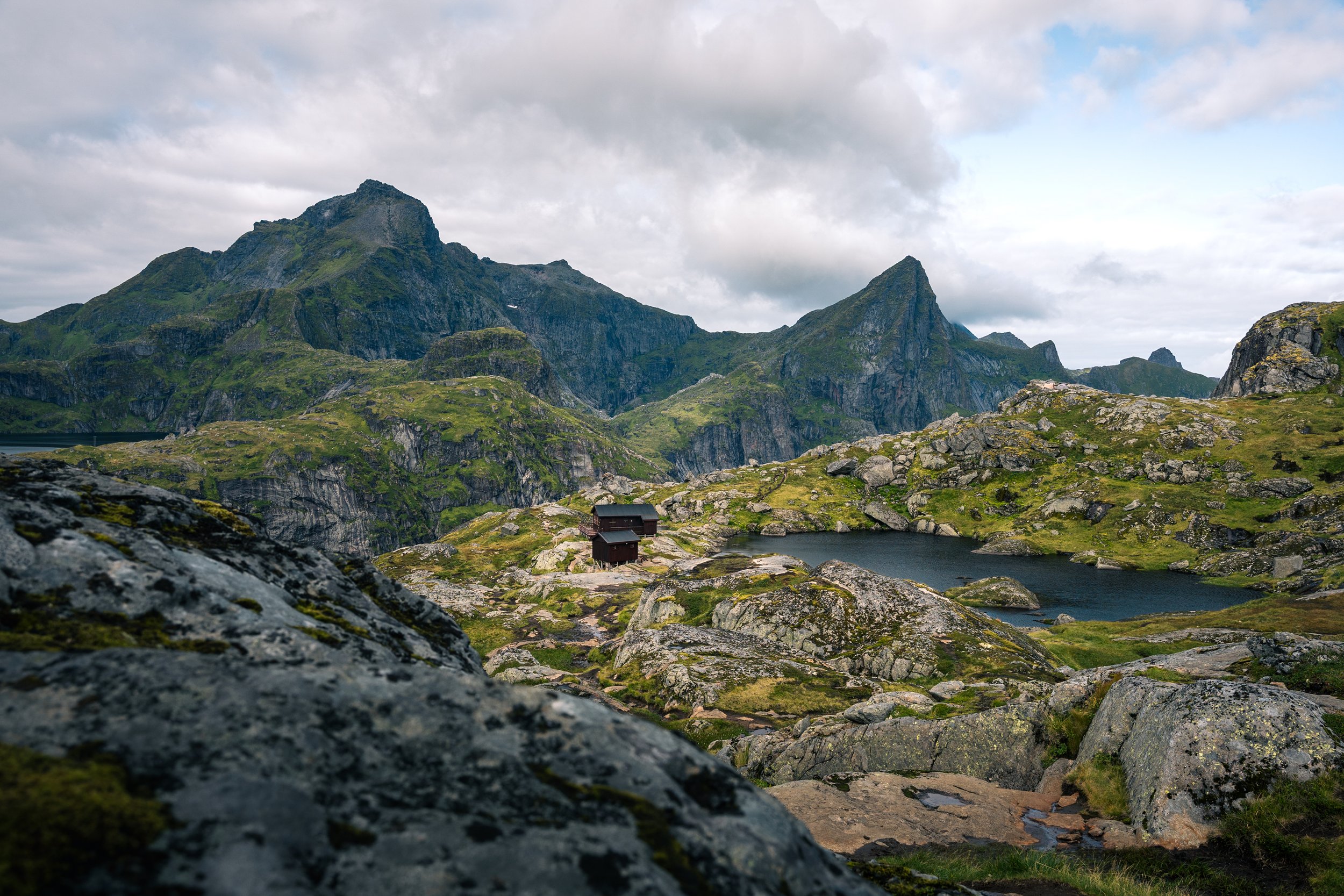 Mountain landscape with green slopes, rocky terrain, a lake, and small houses, under a cloudy sky.