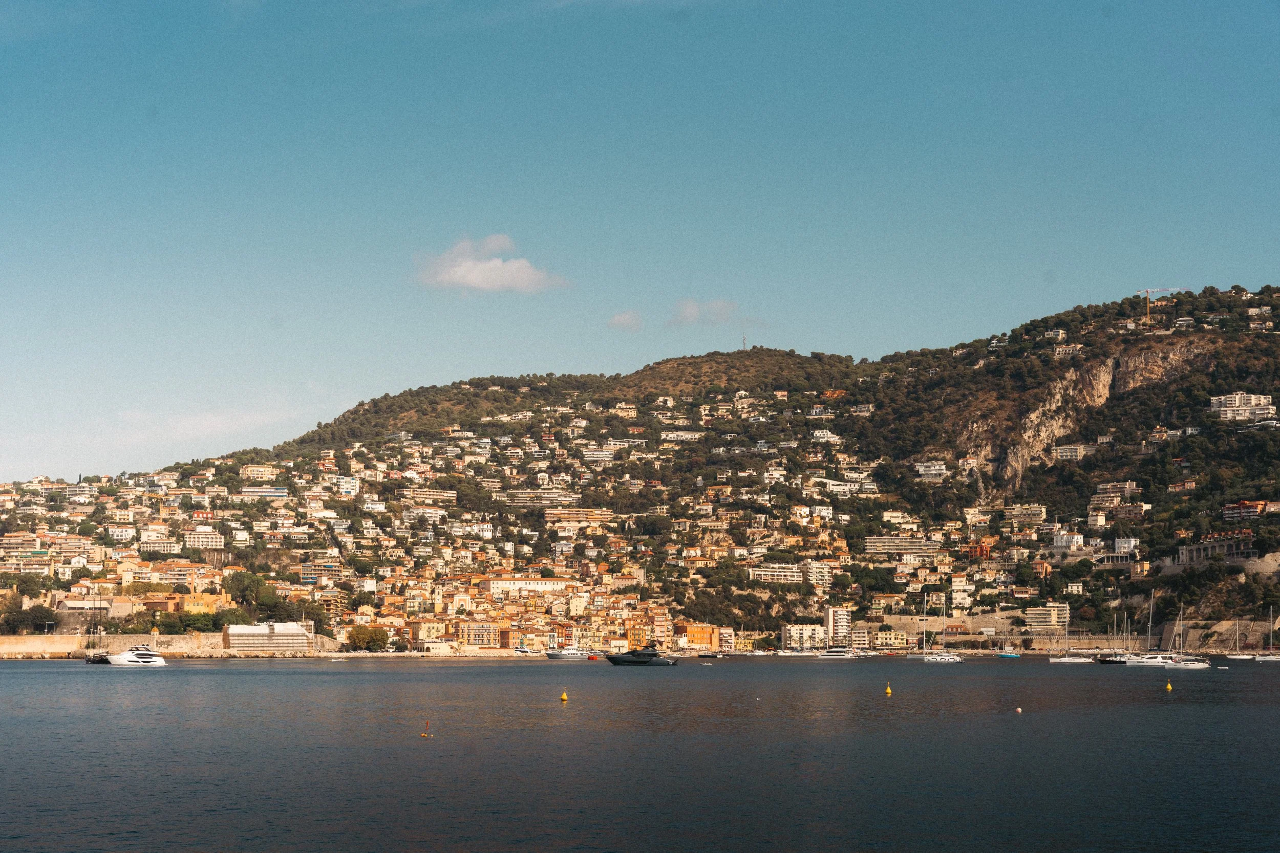 A city hillside with numerous buildings and houses along a slope by the water, with boats and yachts floating on the calm water in the foreground.