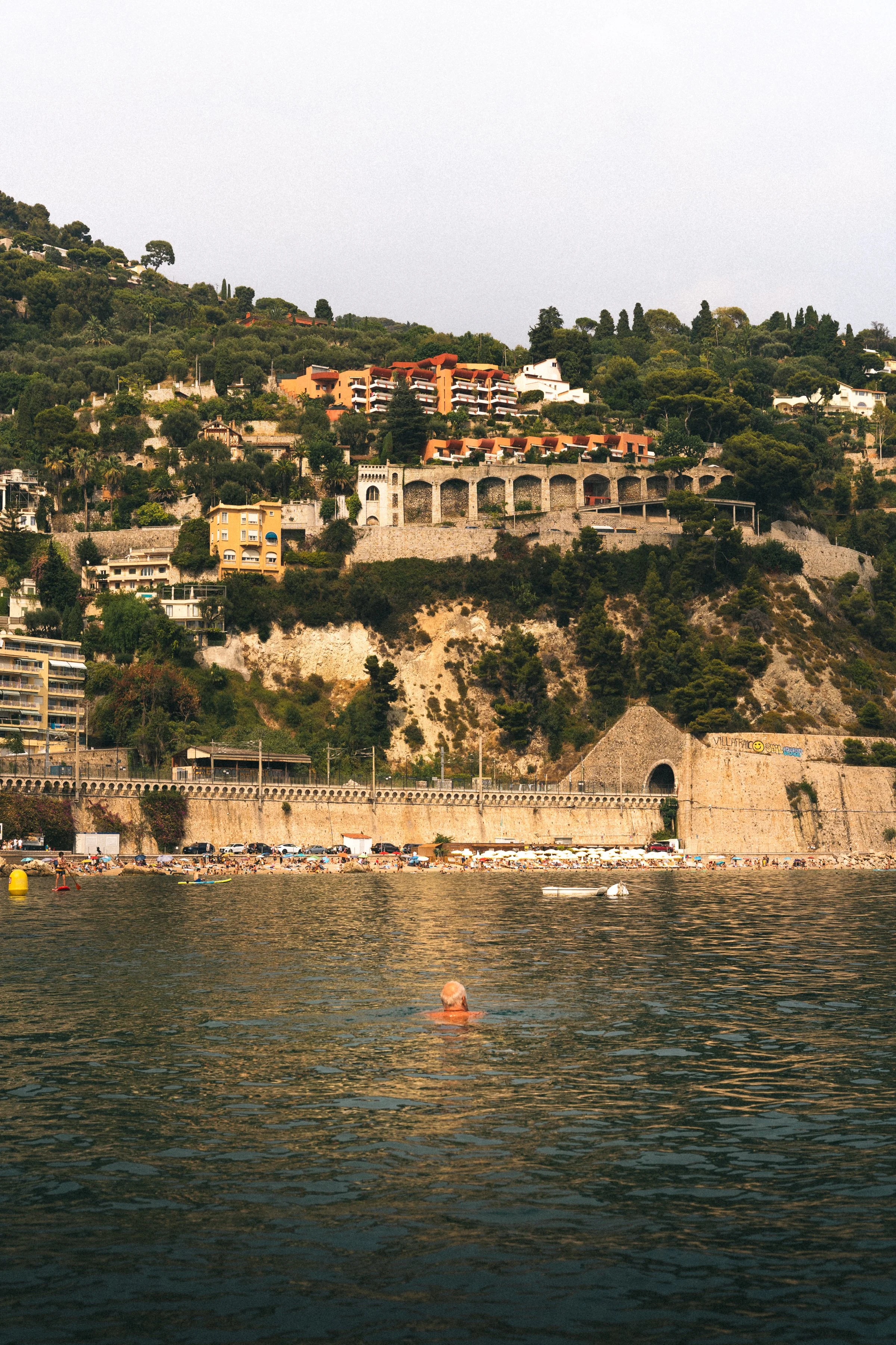 People swimming and lounging at a beach along the water's edge, with hillside houses and greenery in the background.