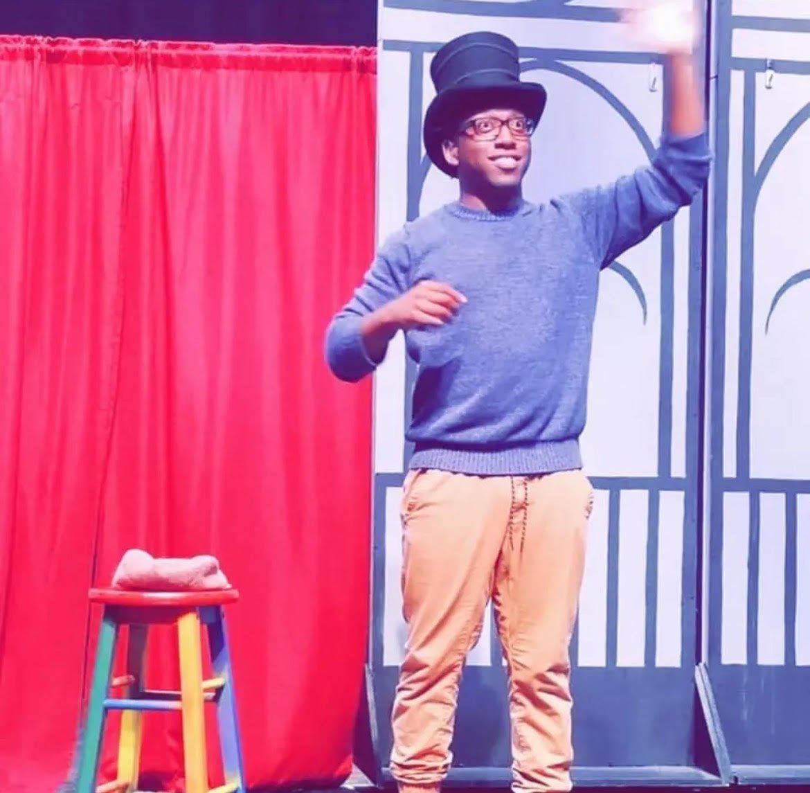 Person standing on stage wearing a top hat, blue sweater, and beige pants, next to a colorful stool. Red curtain and theatrical backdrop in the background.
