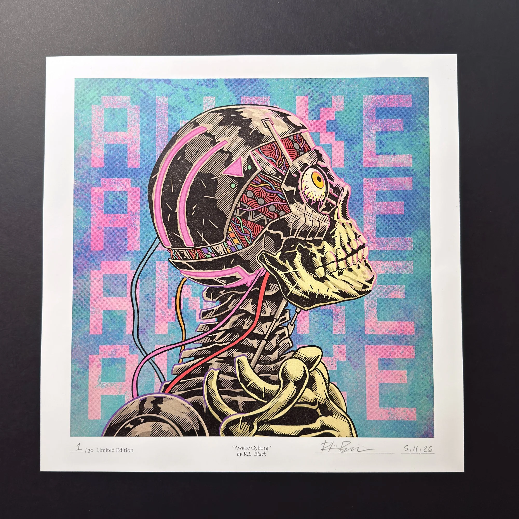 Physical “Awake Cyborg” limited-edition print photographed on a dark surface, showing the full artwork with a white border, hand numbering, artist signature, and date written along the bottom.