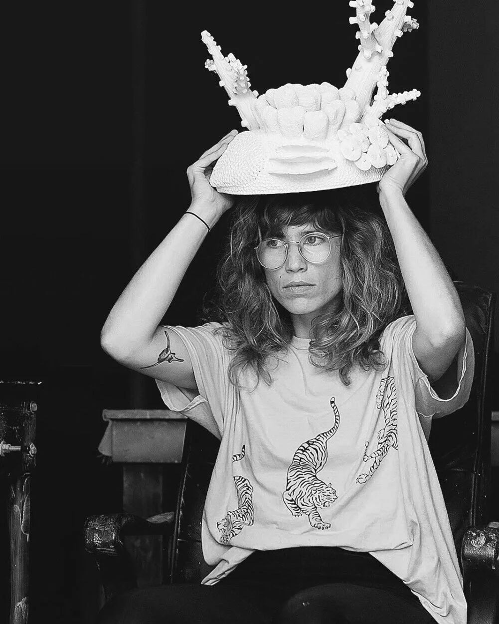 A woman with curly hair and glasses is seated and holding a large, artistic white cake resembling a crown or sculpture.