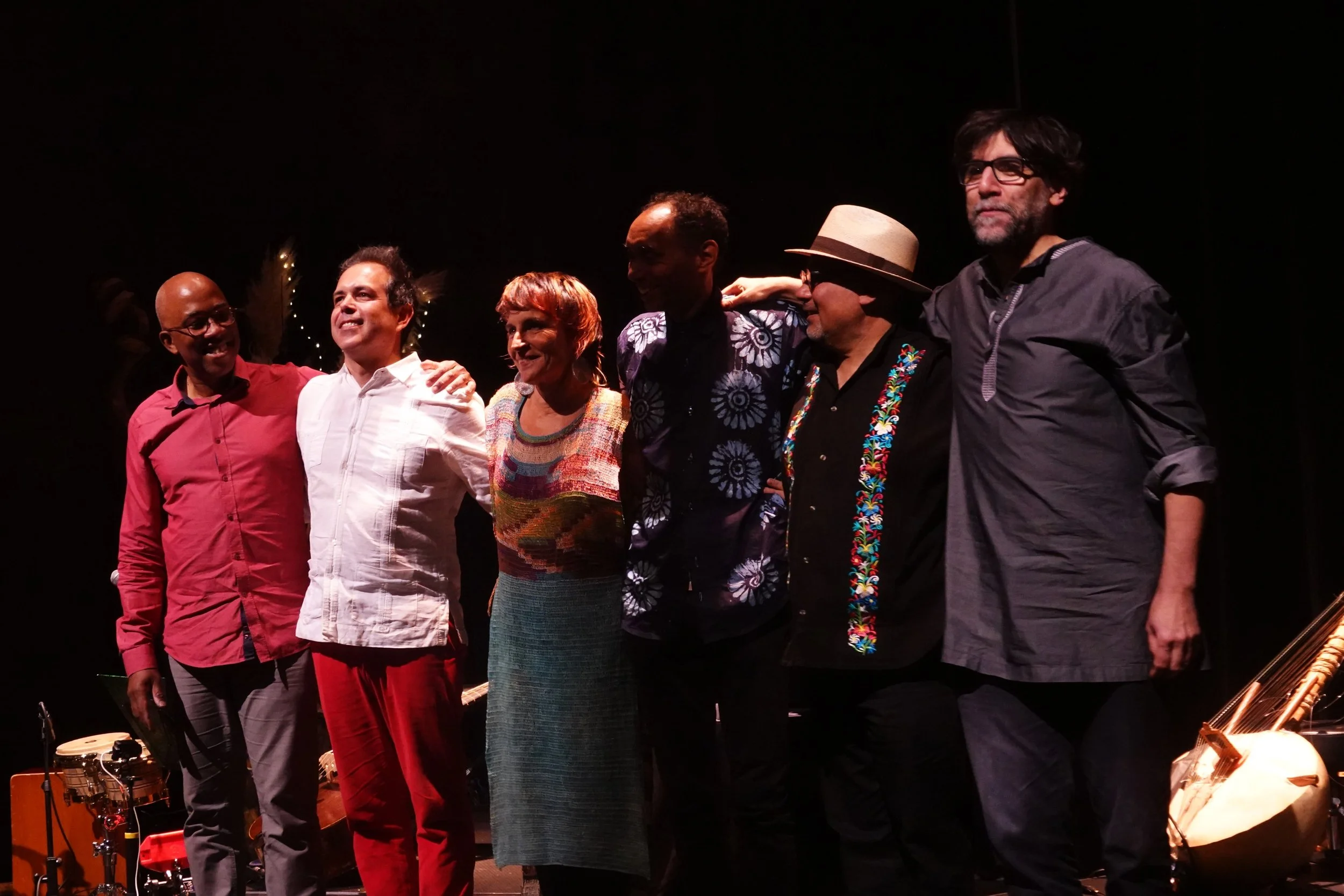 Groupe de sept musiciens ou artistes sur scène, en fin de performance, applaudissant et se saluant, avec instruments de musique visibles, dans une ambiance chaleureuse et conviviale.