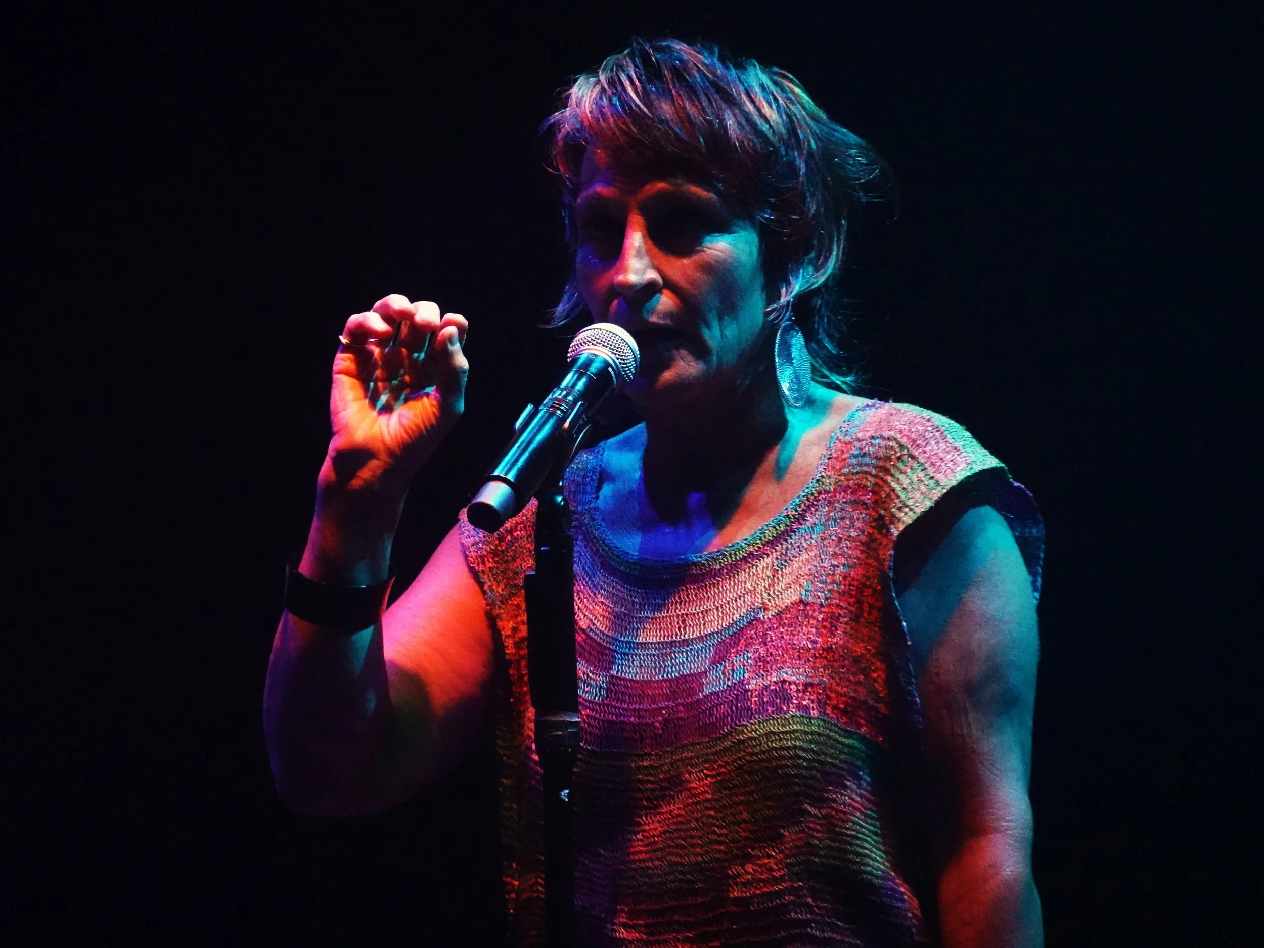 Une femme chante dans un micro sous un éclairage coloré sombre, portant un haut multicolore et un grand boucles d'oreilles