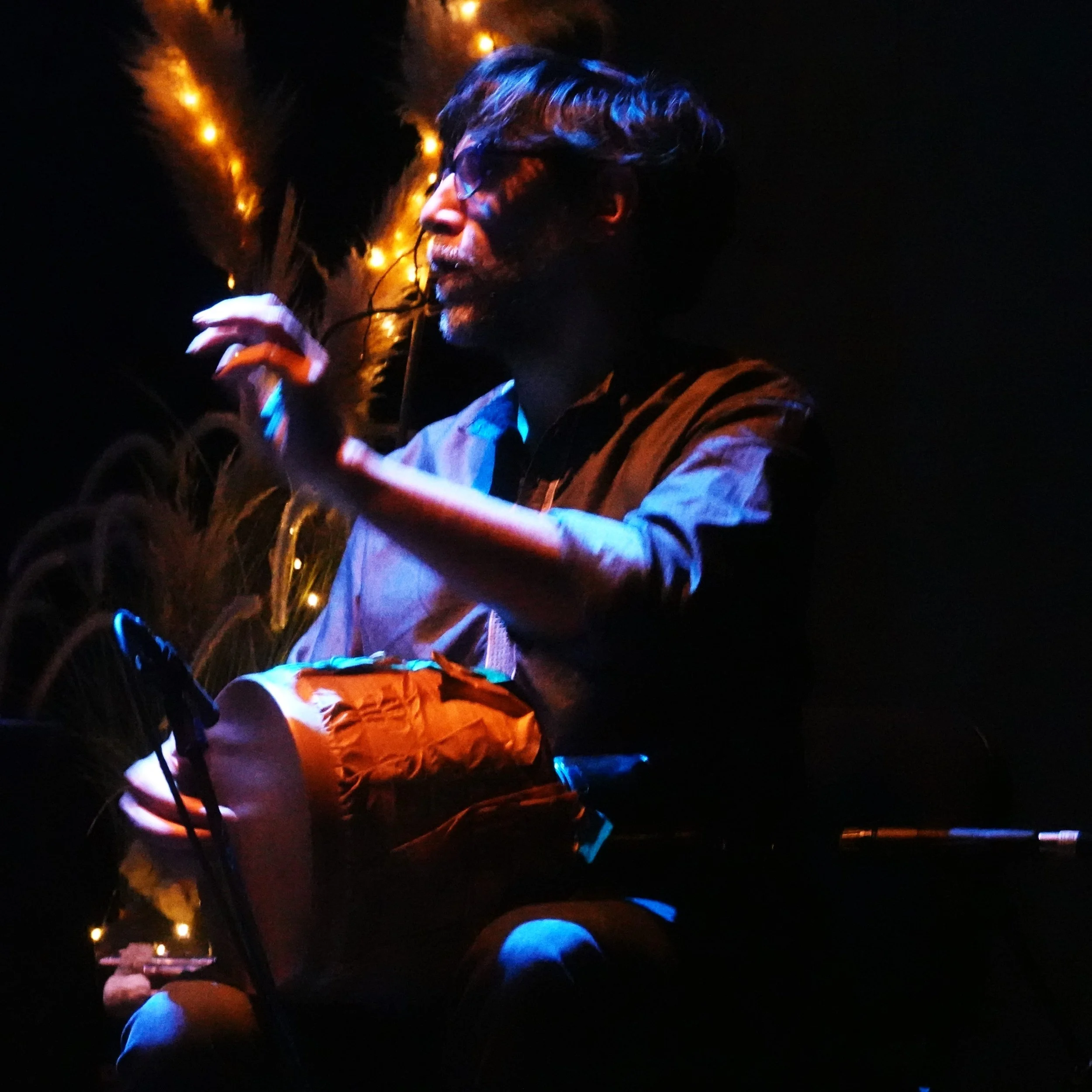 Un homme avec des cheveux longs portant des lunettes, éclairé par des lumières chaudes dans l'obscurité, tenant un objet dans sa main.