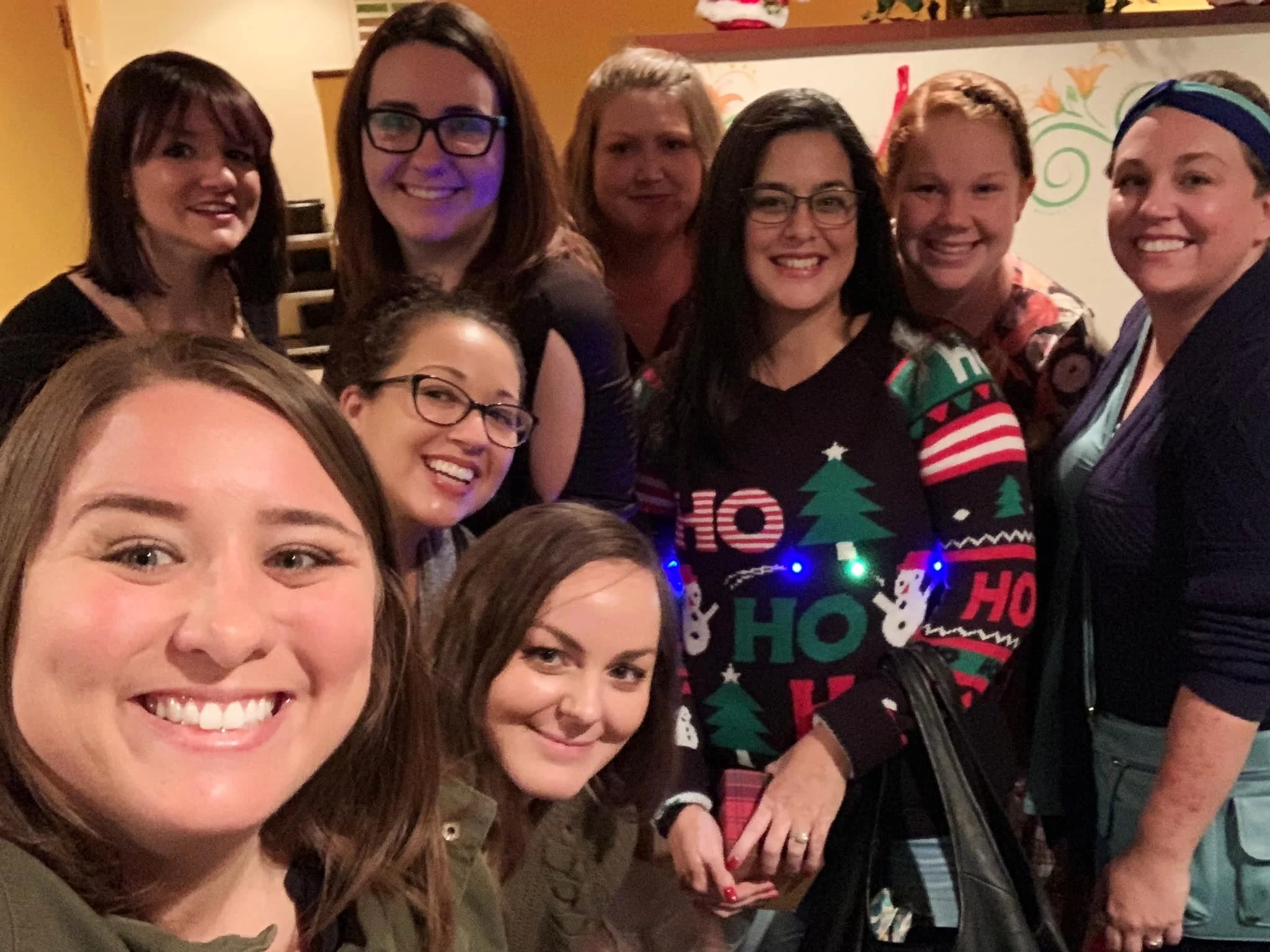 A group of smiling women posing for a photo at a festive gathering, one wearing a Christmas sweater.