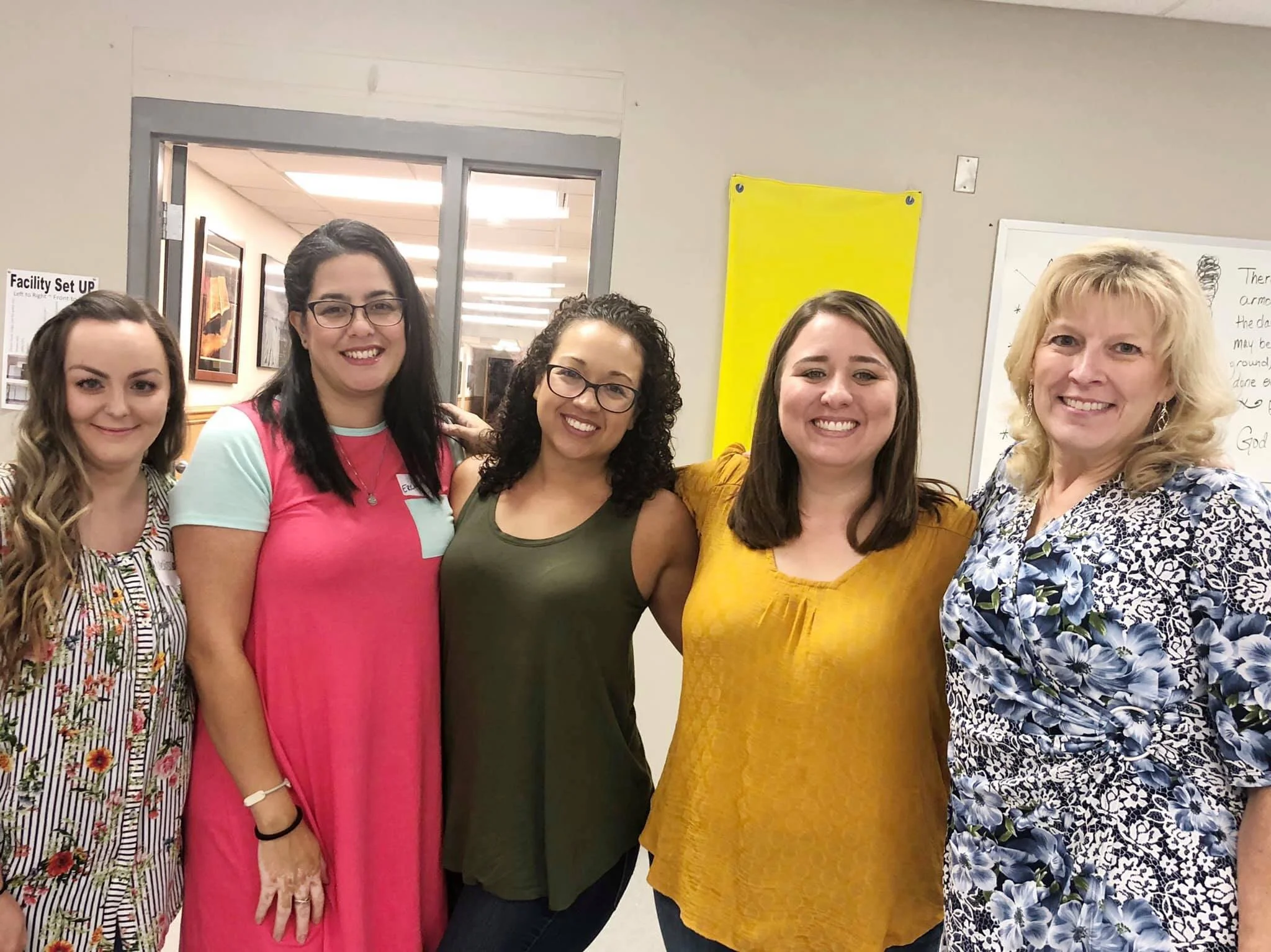 Group of five women standing together in an indoor setting, smiling at the camera. They are dressed in colorful and patterned clothing, with a whiteboard and a yellow bulletin board visible in the background.
