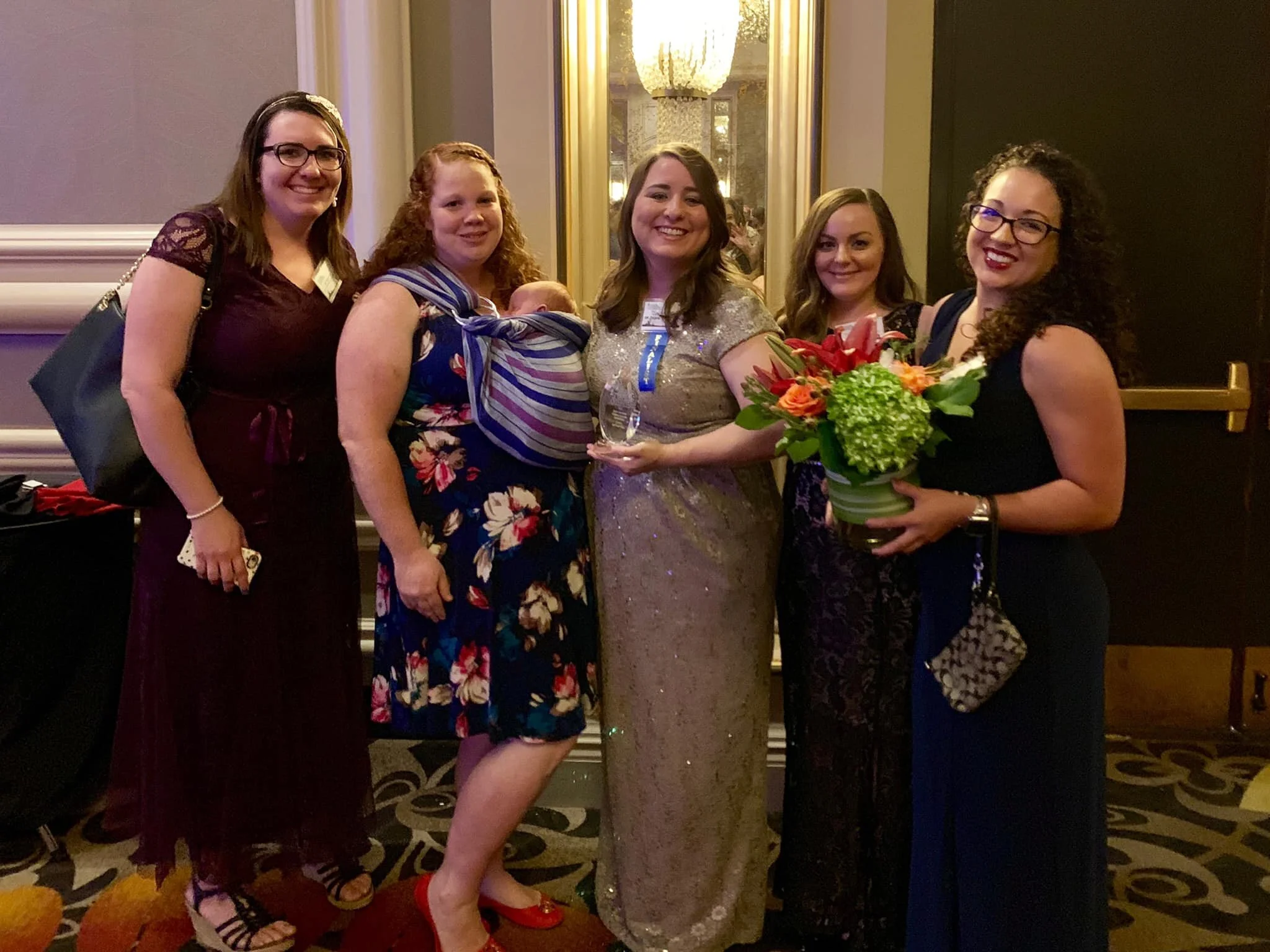 A group of five women, including a woman holding a baby and a woman holding a bouquet of flowers, standing in a line at an indoor event with a chandelier and mirror in the background.