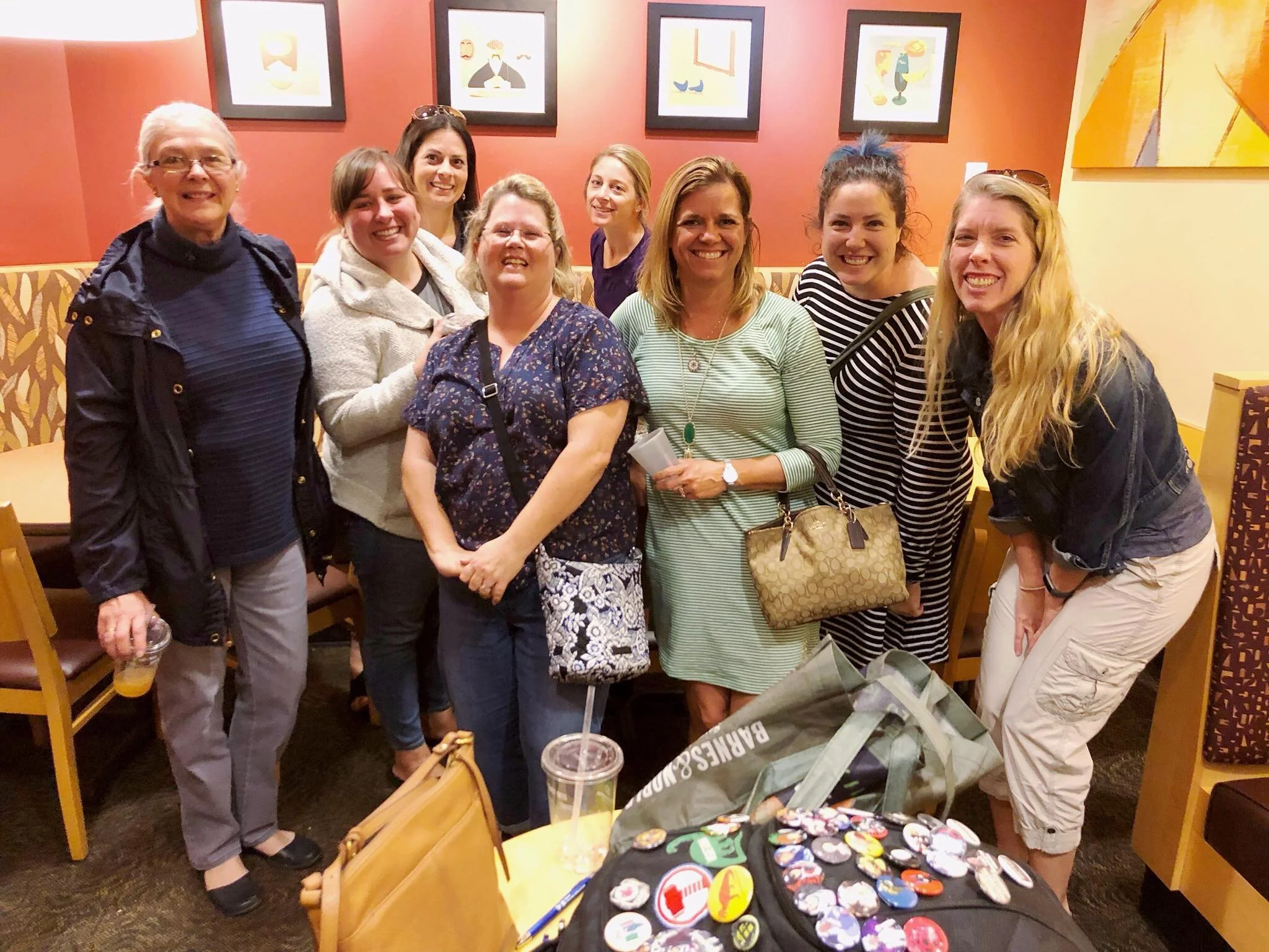 Group of nine women smiling in a restaurant, standing behind a table with various pins and badges, some wearing glasses, and colorful wall art in the background.