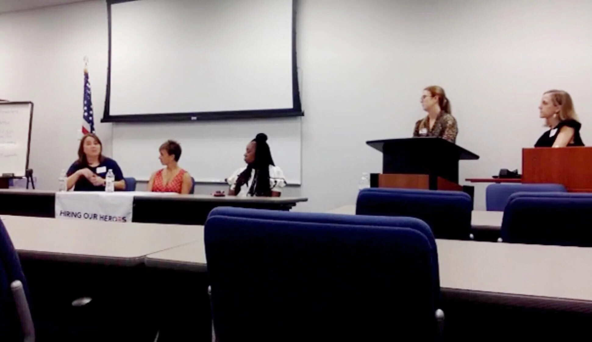 Four women in a room with chairs and tables, one woman standing at a podium, while third woman sits at a table near a whiteboard and with a water bottle, and a woman in a black top sitting at the far right, behind a desk with another water bottle. Th