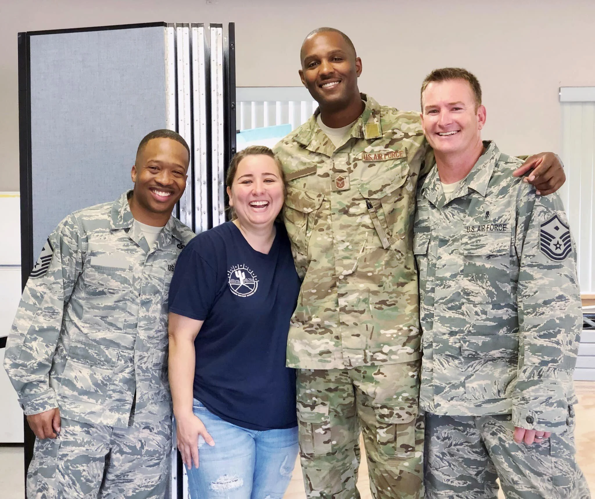 Four people, two men in U.S. Air Force uniforms, a woman, and a man in casual clothing, smiling and posing together indoors, with a folding divider and window blinds in the background.