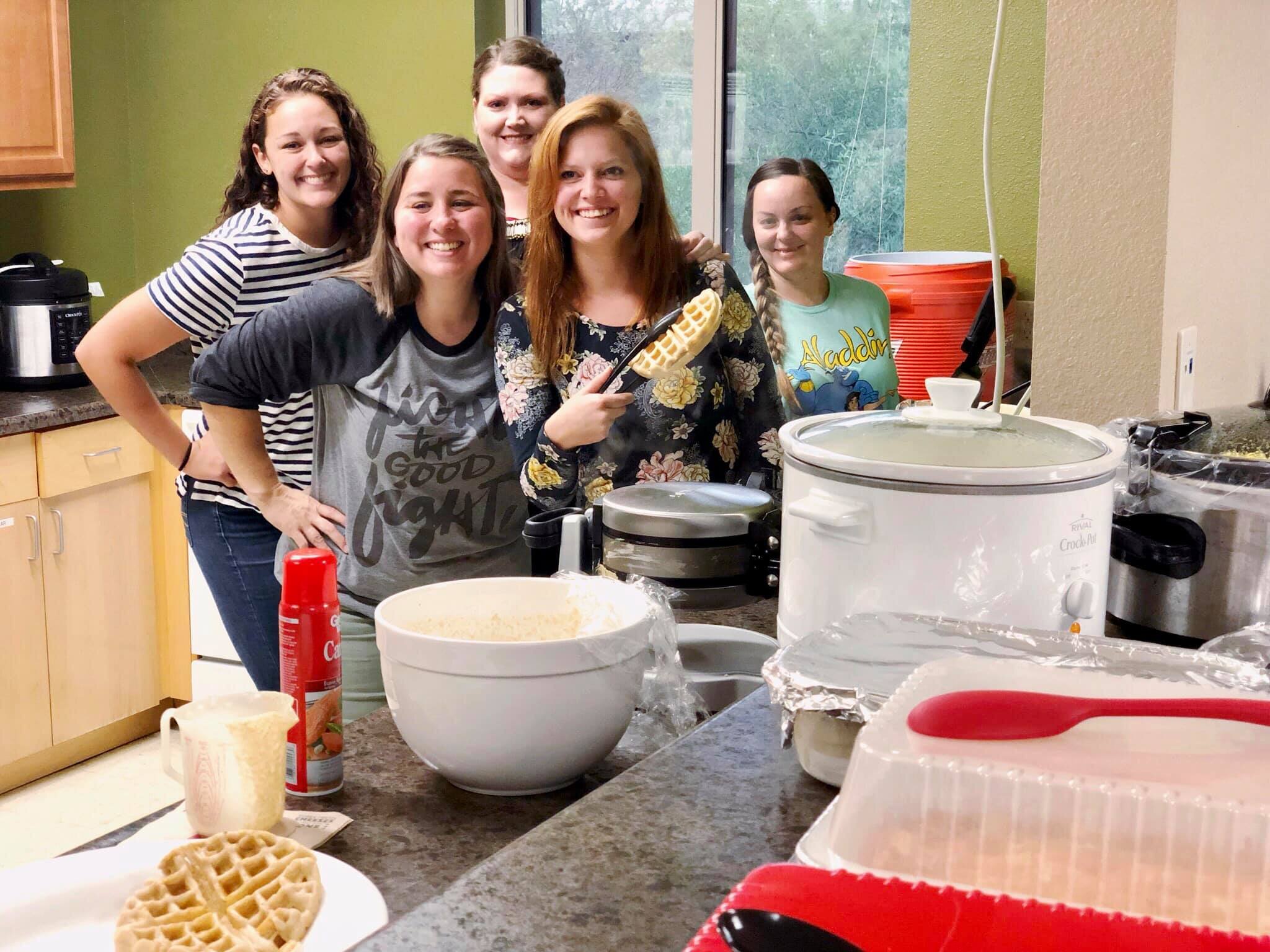 A group of six women in a kitchen smiling at the camera, with baking items and waffle cones on the counter, in front of a large window.