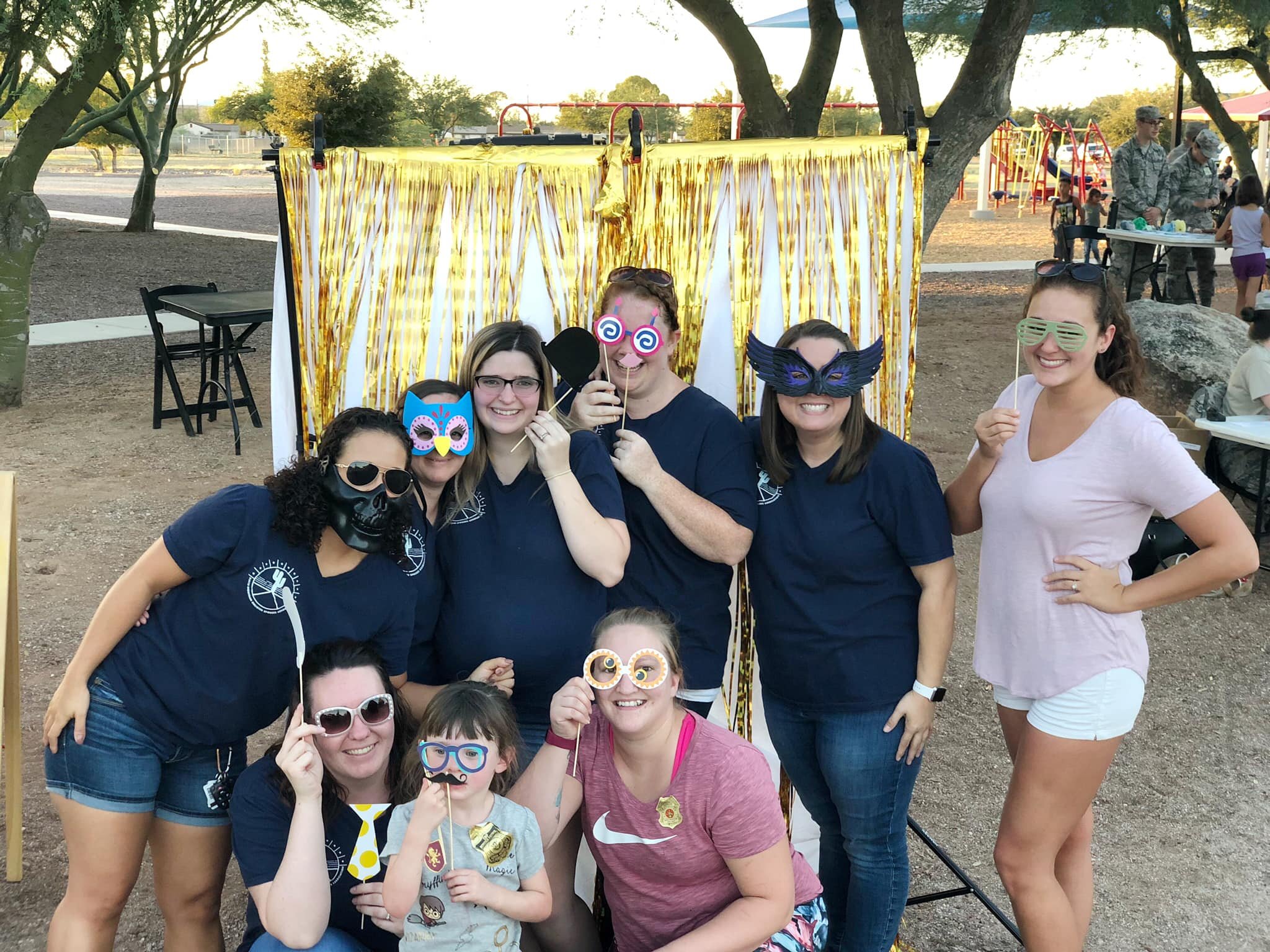 A group of smiling women and children at a park, some wearing fun masks and costumes, standing in front of a gold and white photo booth backdrop with trees in the background.