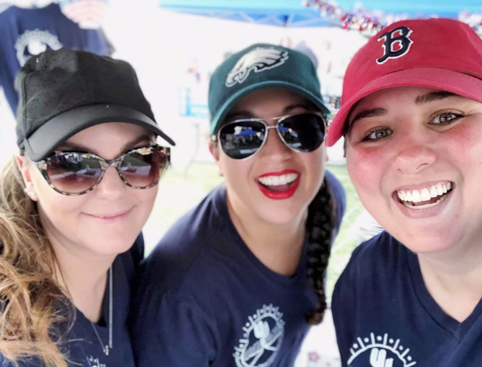Three women smiling at a gathering, wearing baseball caps and sunglasses, with a cheerful outdoor background.
