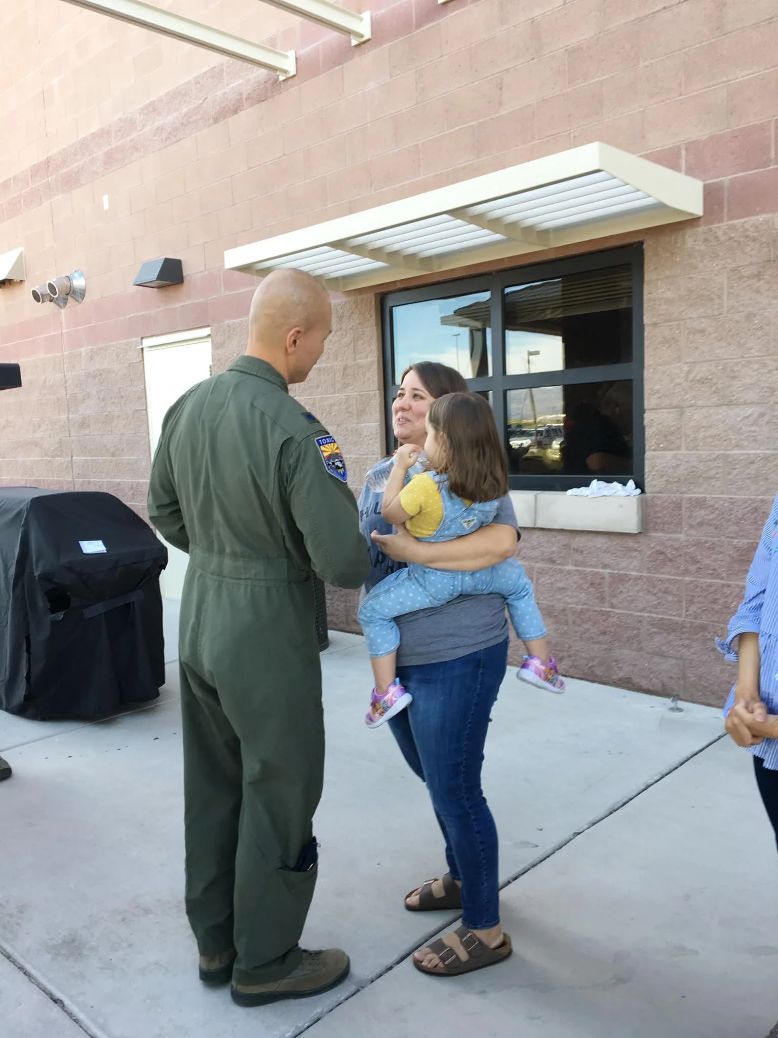 A woman holding a young girl in her arms, facing a man in a military uniform, outside a building with a brick wall and window.