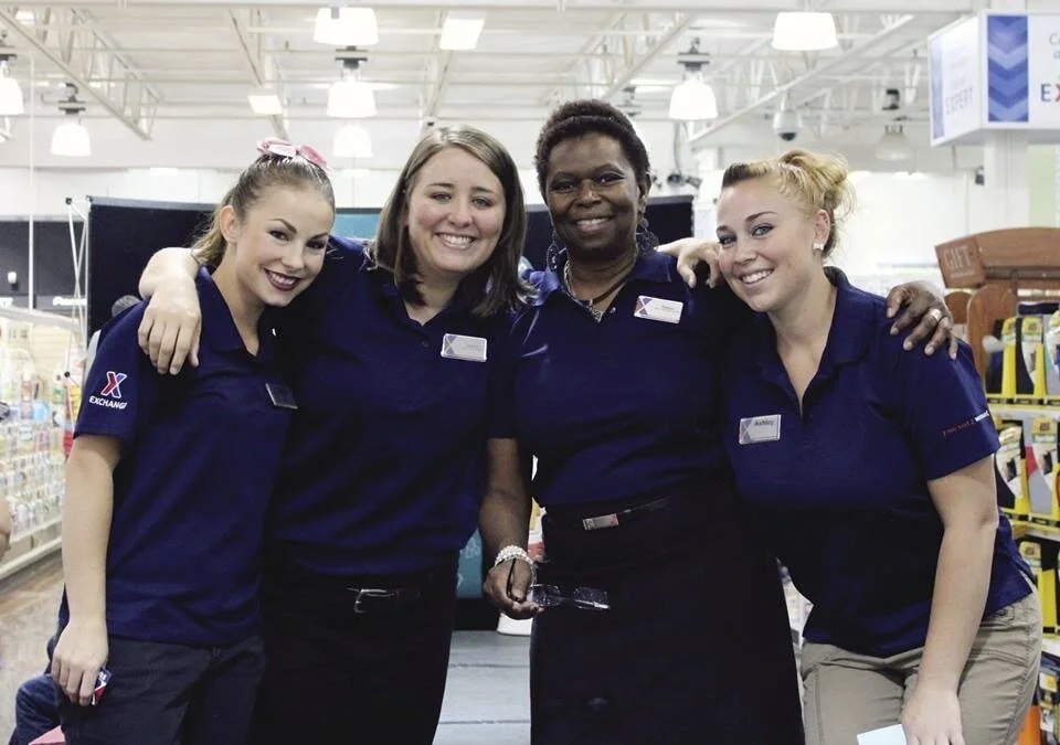 Four women in blue uniforms smiling and hugging in a store aisle.
