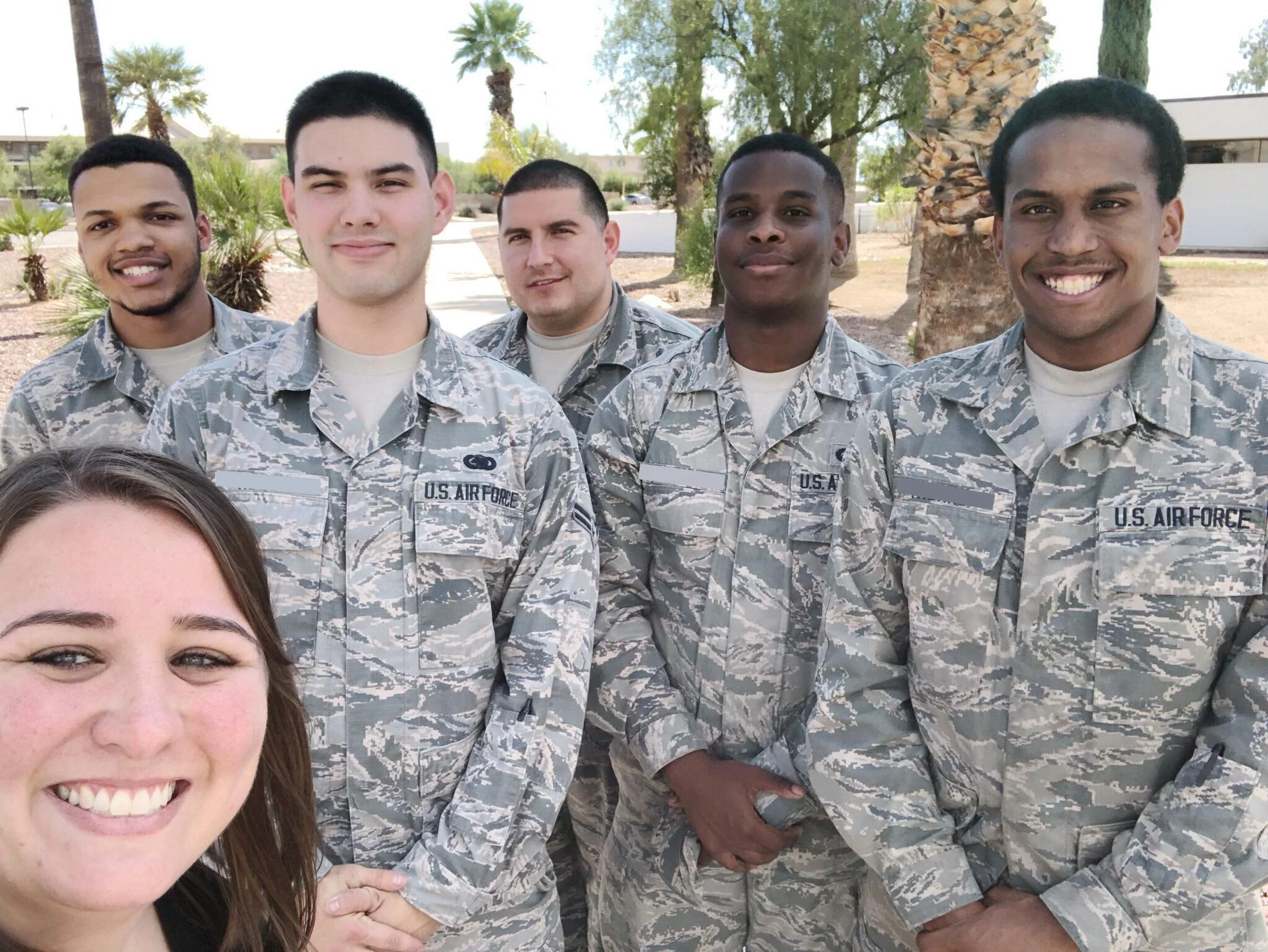 Group photo of five smiling military personnel in camouflage uniforms outdoors with palm trees and a building in the background.