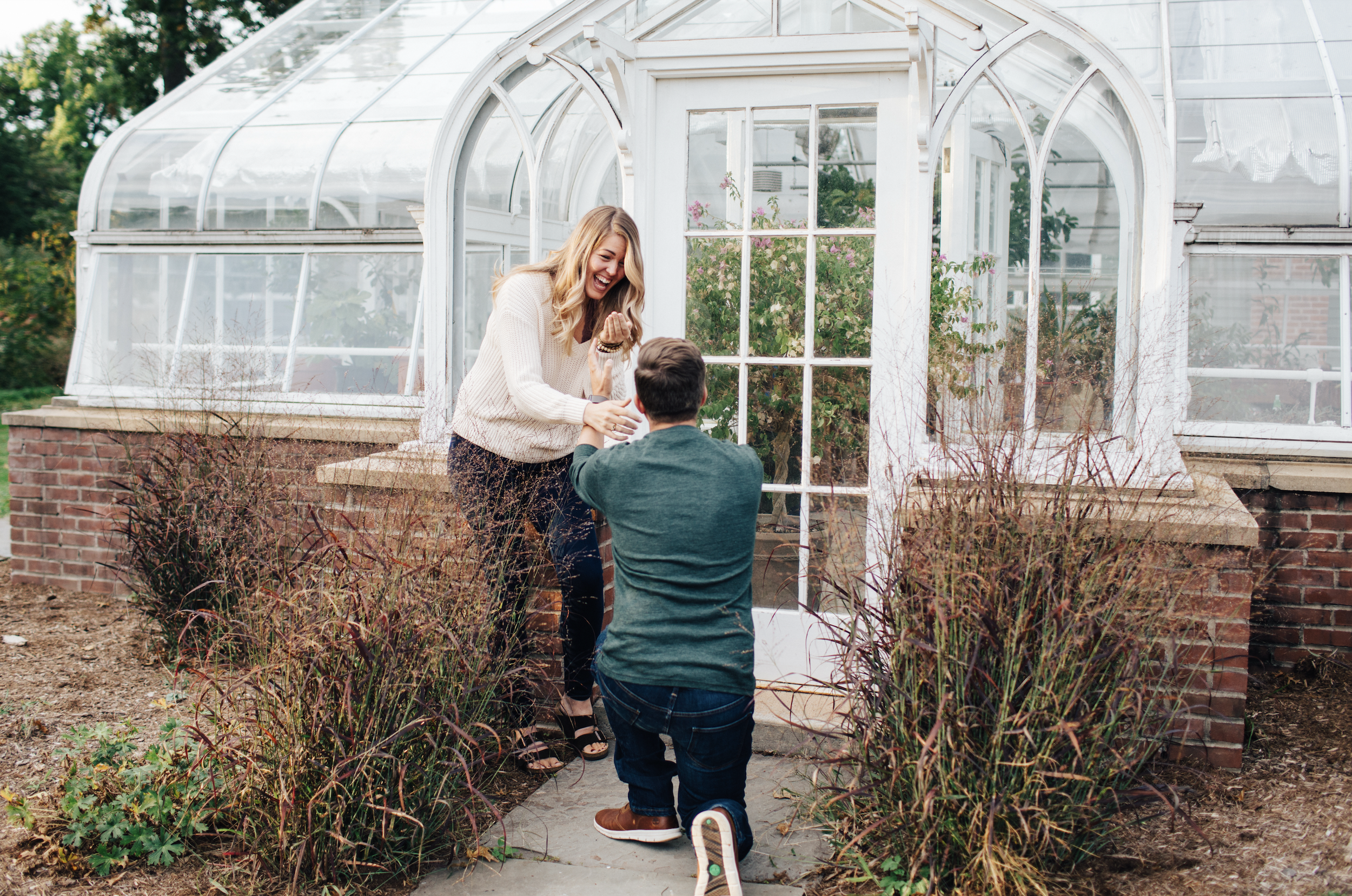 A couple is in front of a greenhouse. The woman is standing and laughing, while the man is kneeling and holding her hand, proposing marriage.