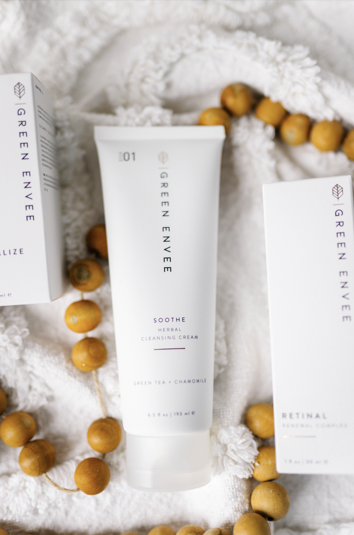 Green Envee Soothe Herbal Cleansing Cream in a white tube on a white fabric with wooden bead garland.