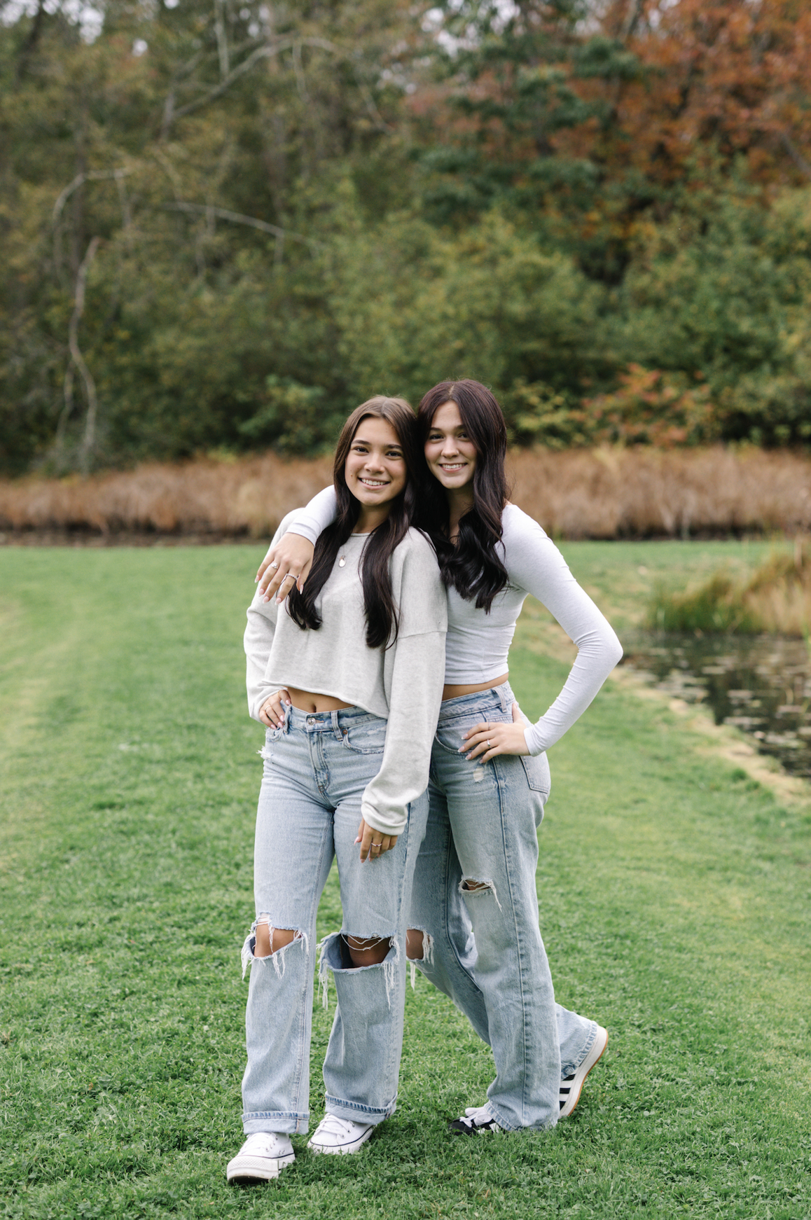 Two young women with dark hair standing close together outdoors by a pond, smiling, with autumn foliage in the background.