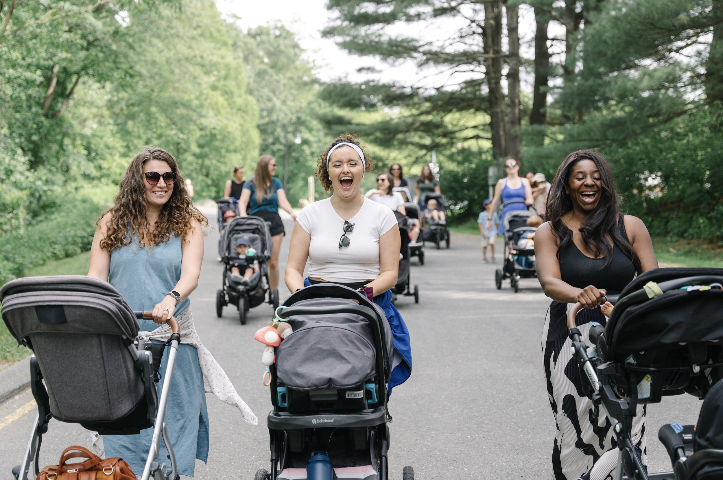 Three women walking with strollers in a park with green trees, smiling and enjoying a sunny day.