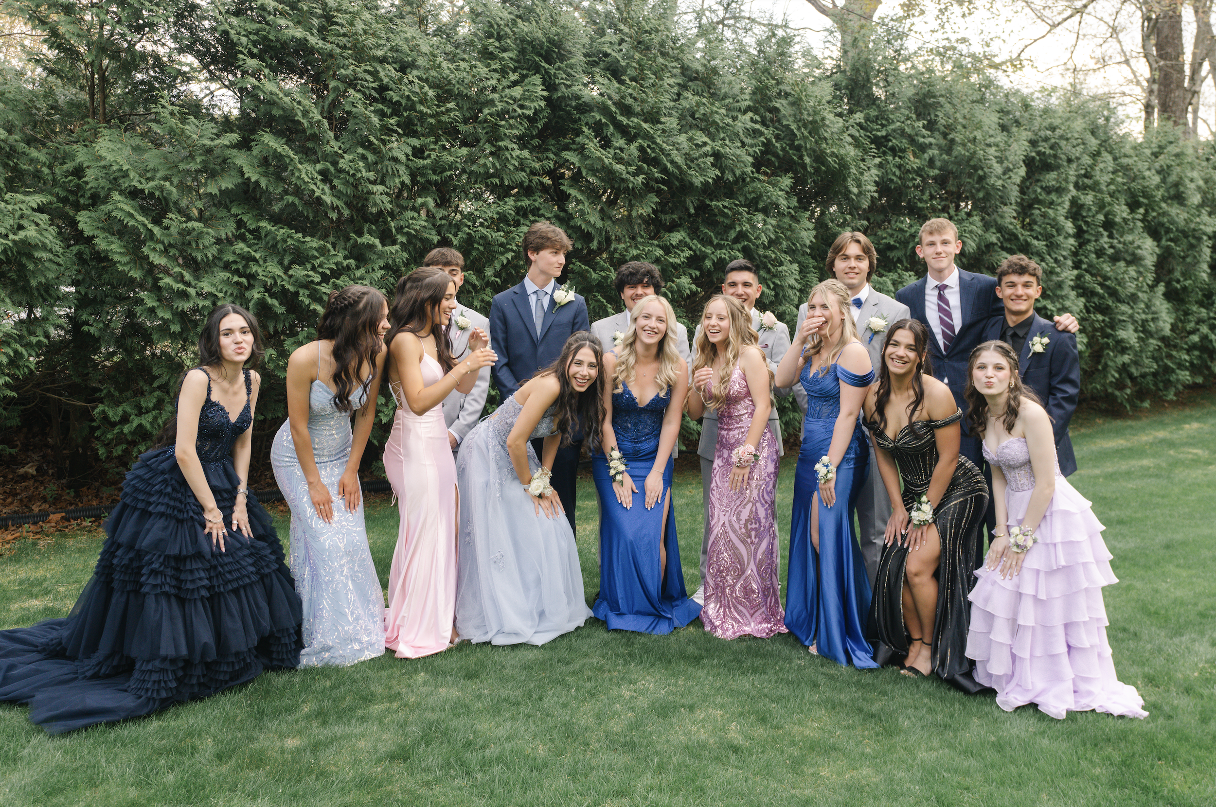 Group of teenagers dressed in formal prom attire, standing on a grassy lawn in front of dense green bushes, smiling and laughing.