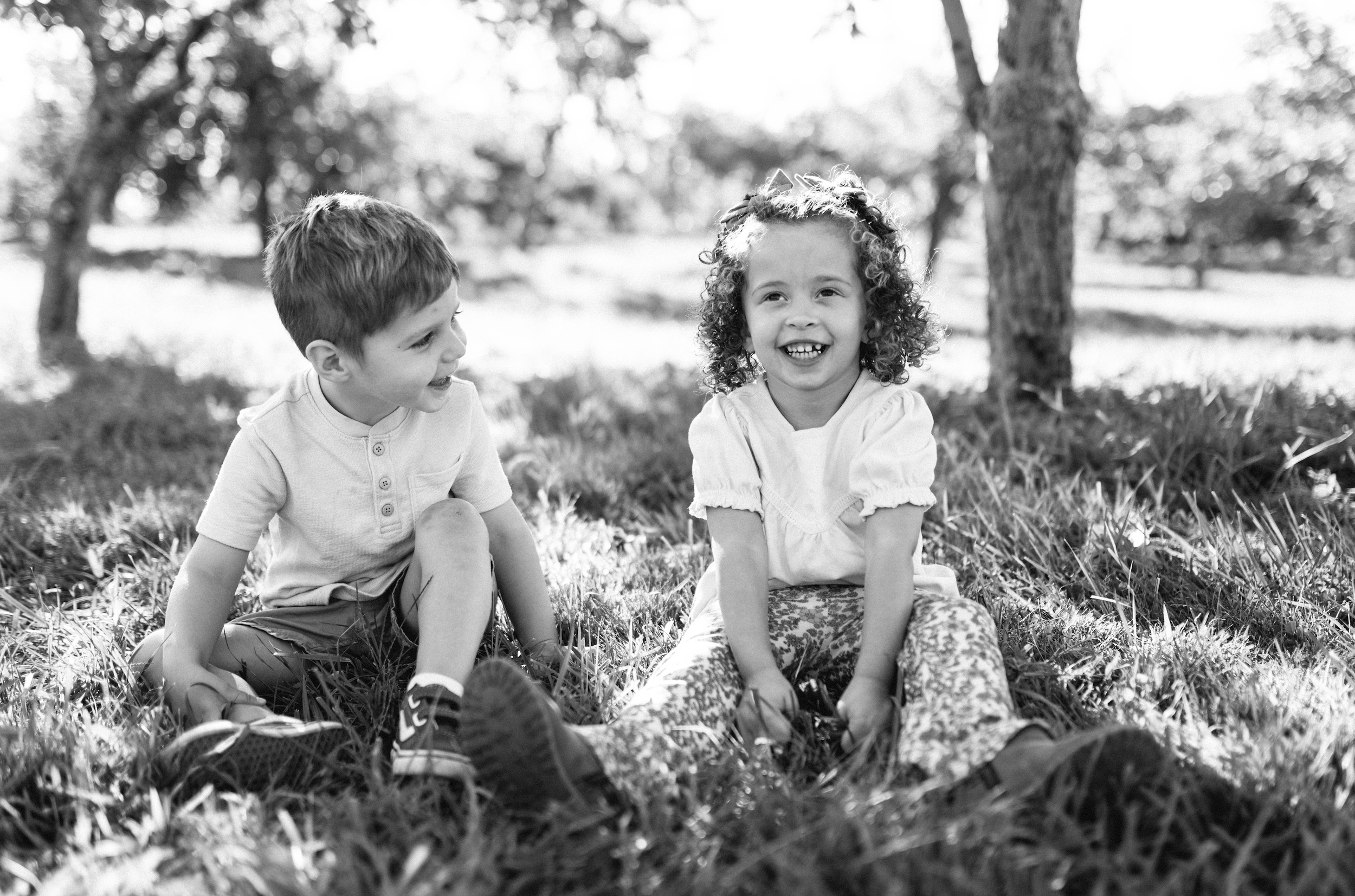 Two children, a boy and a girl, sitting on the grass outdoors, smiling and laughing together.