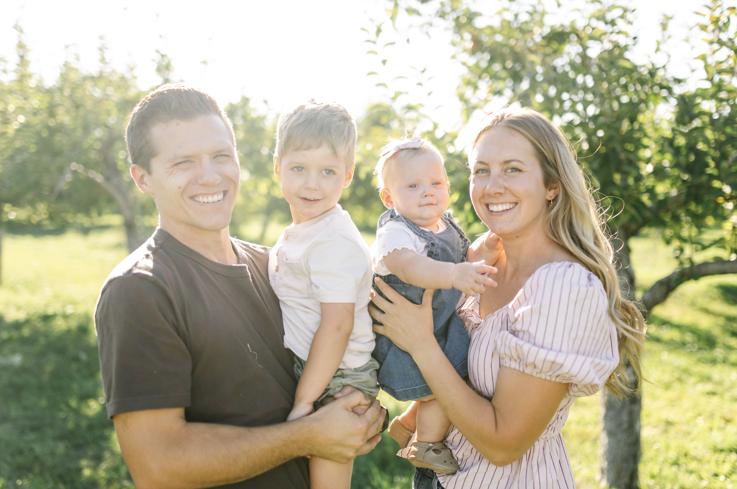 Happy family of four in an orchard on a sunny day, with a young man and woman each holding a small child.