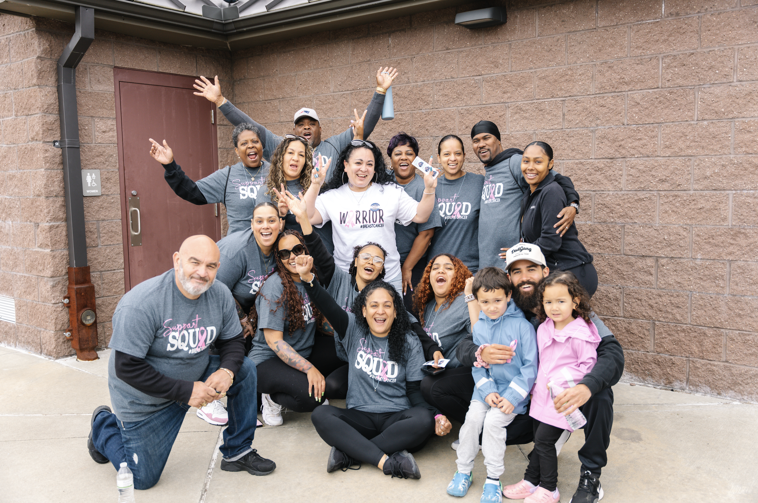 Group of adults and children smiling and celebrating outdoors in front of a brick building, some wearing matching gray T-shirts with 