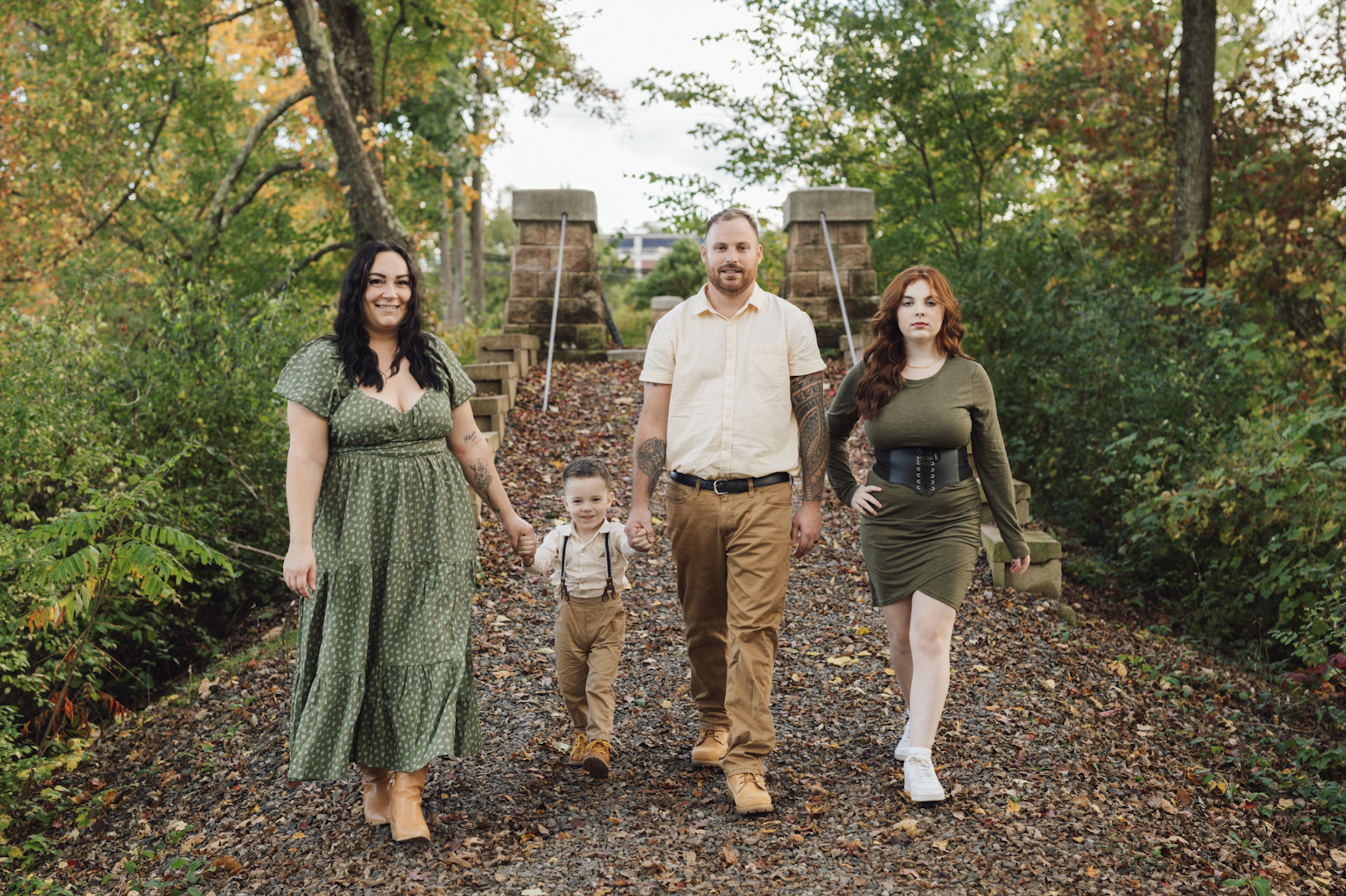 A family of five walking hand-in-hand on a wooded trail during autumn. The woman on the left has black hair and wears a green polka-dot dress. The young boy next to her has short hair, wearing a beige shirt, khaki pants, and suspenders. The man in the center has a beard, tattoos, and wears a beige shirt with khaki pants. The woman on the right has long red hair, wears a green dress with a black belt, and white sneakers. They are walking on a leaf-covered path with trees and a small stone bridge in the background.