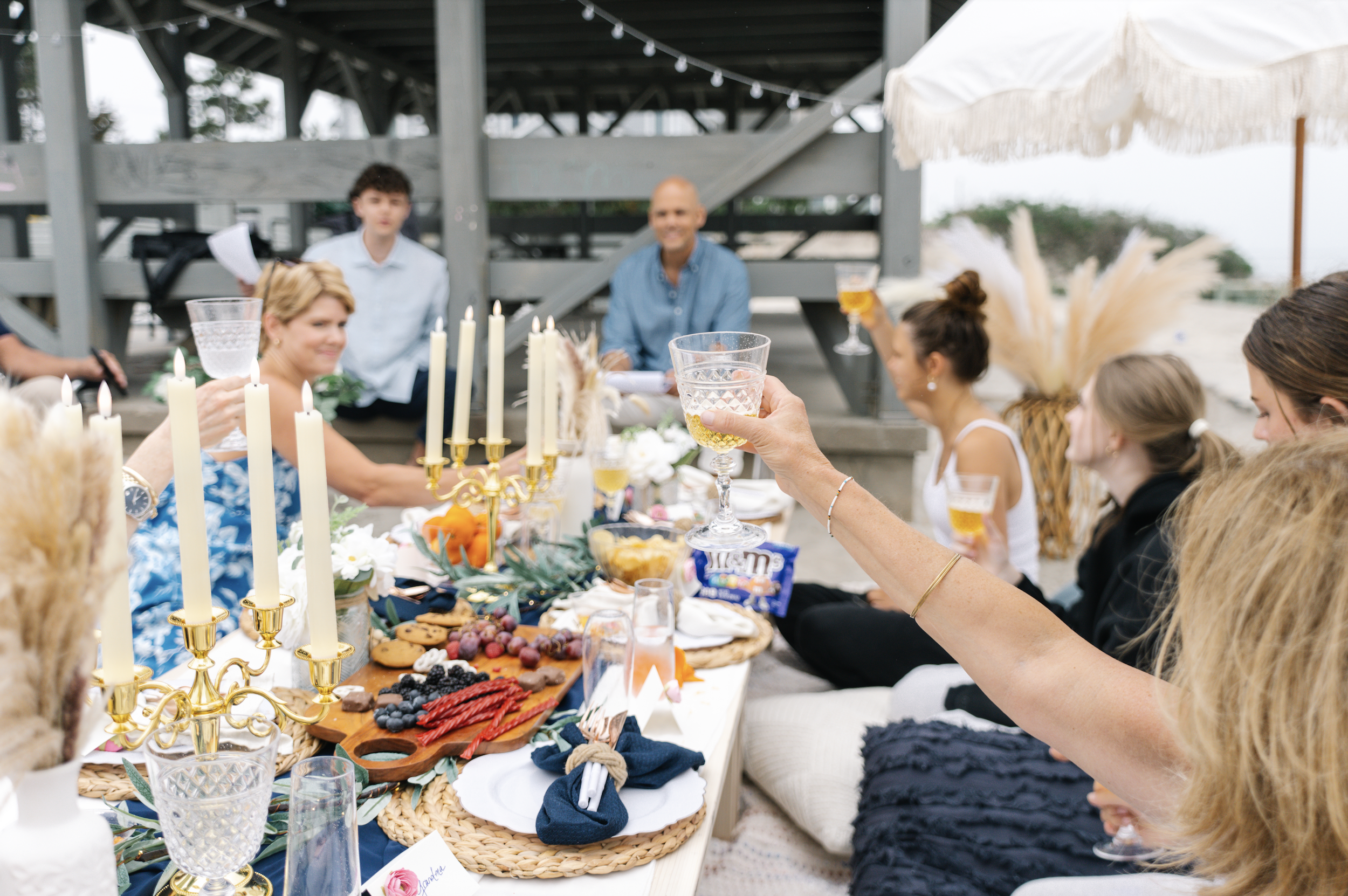 People gathered around a decorated outdoor table for a celebration, raising glasses in a toast. The table is adorned with candles, flowers, and food, with a beach and overcast sky in the background.