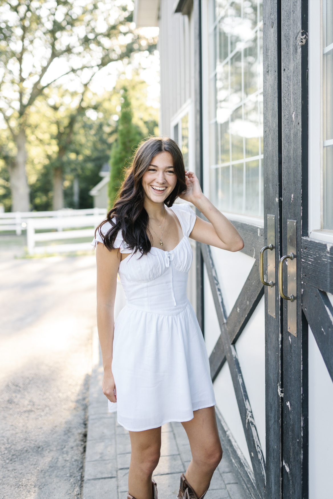 Young woman with long dark hair smiling in a white dress near a barn with large windows