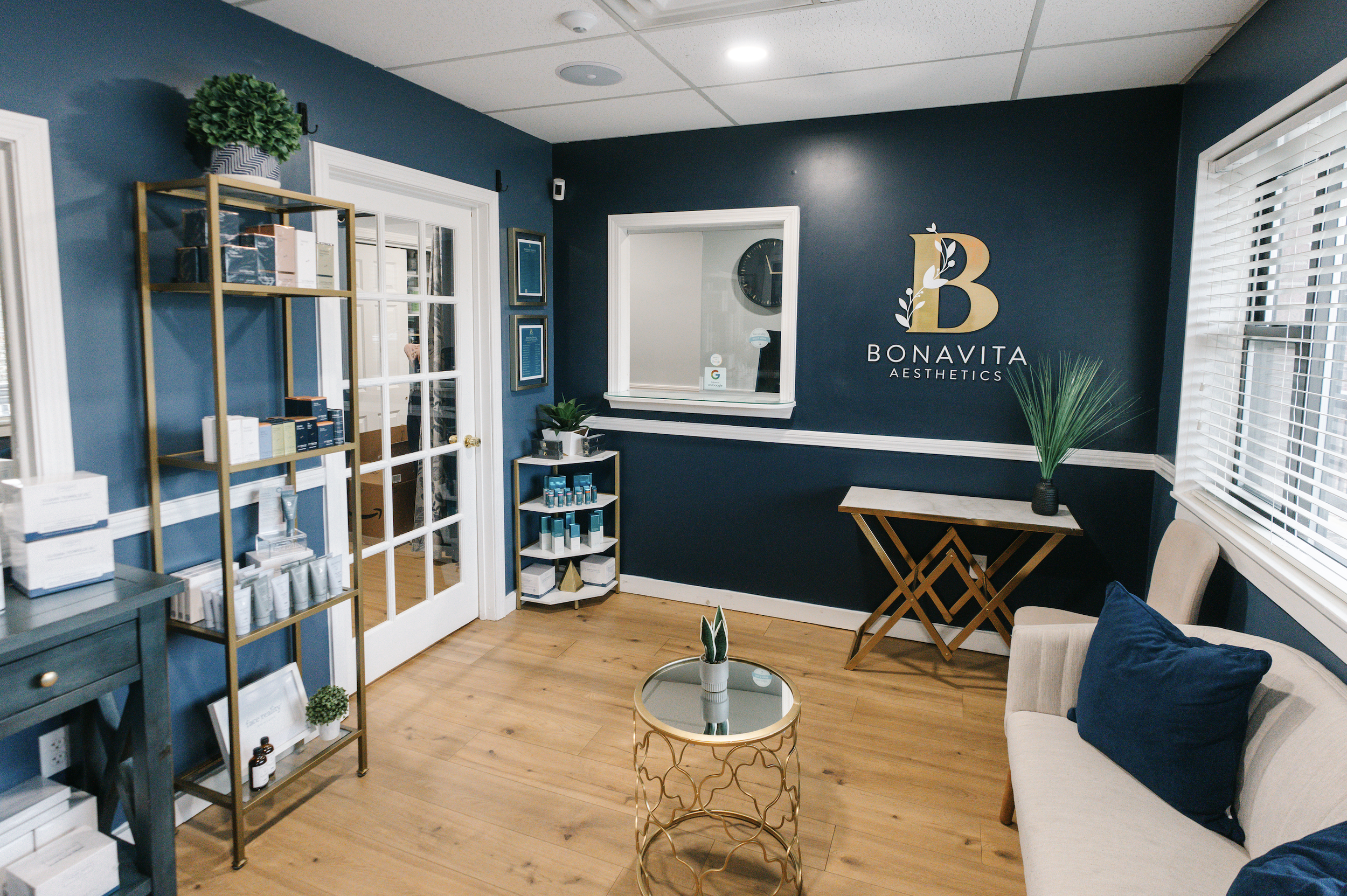 Interior of Bonavita Aesthetics with blue walls, wooden flooring, a beige sofa with navy pillows, a glass-top round coffee table with a potted plant, a gold shelving unit with beauty products, and a window with white blinds.