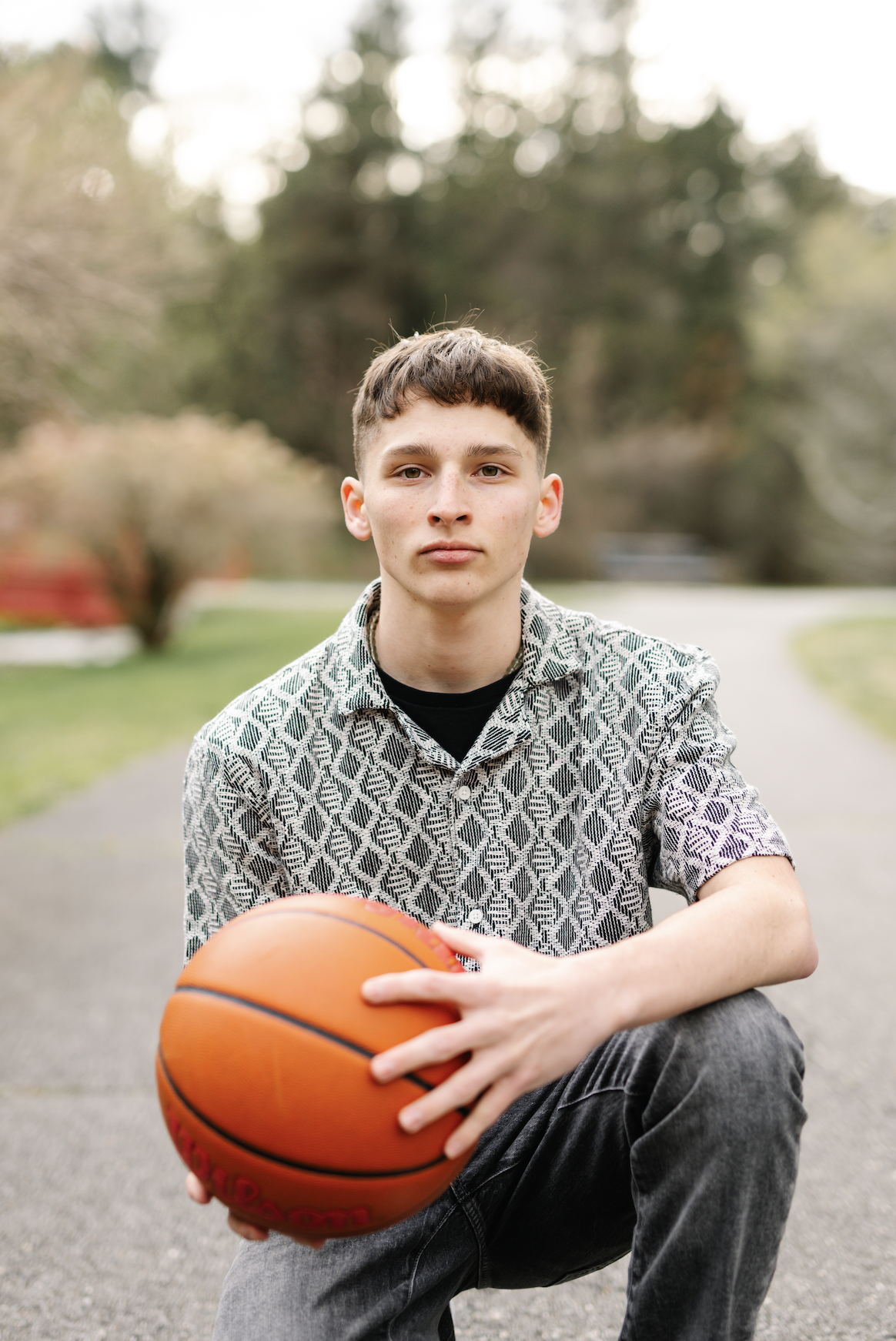 A young man with short brown hair and fair skin holding an orange basketball outdoors on a paved path, with trees and greenery in the background.