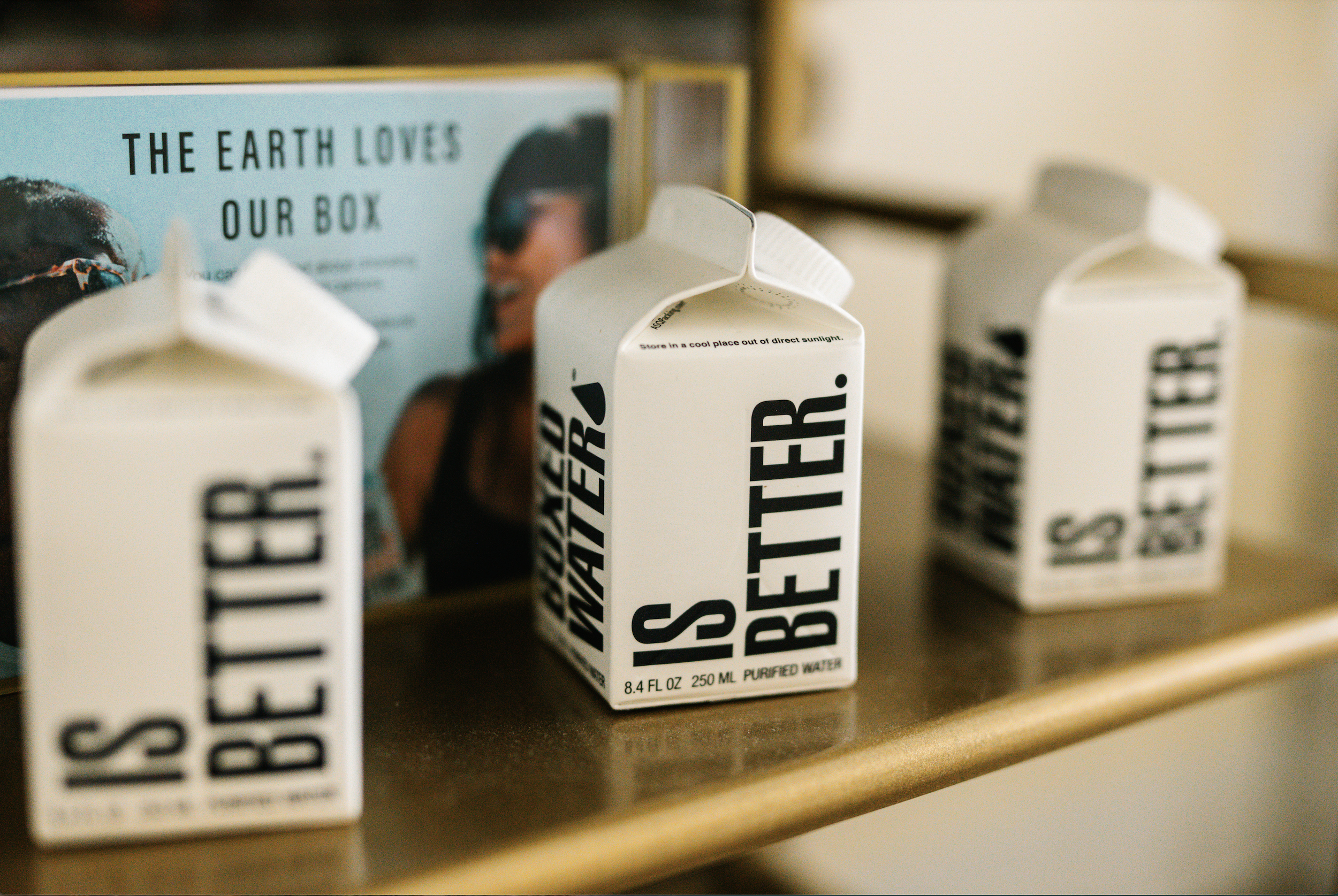 Three cartons of water labeled 'S WATER' on a wooden shelf with a framed poster behind them that says 'THE EARTH LOVES OUR BOX' and features a woman smiling.