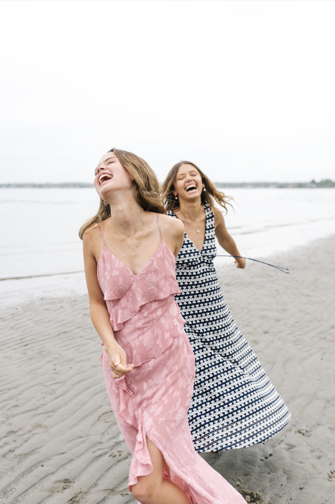 Two women wearing summer dresses laughing and running on a beach under a cloudy sky.
