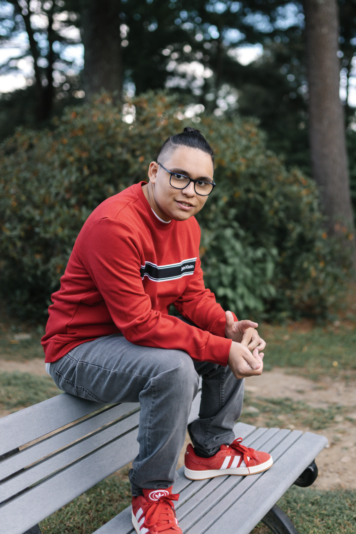 Young man with glasses, wearing a red sweatshirt and gray pants, sitting on a park bench surrounded by trees and bushes.
