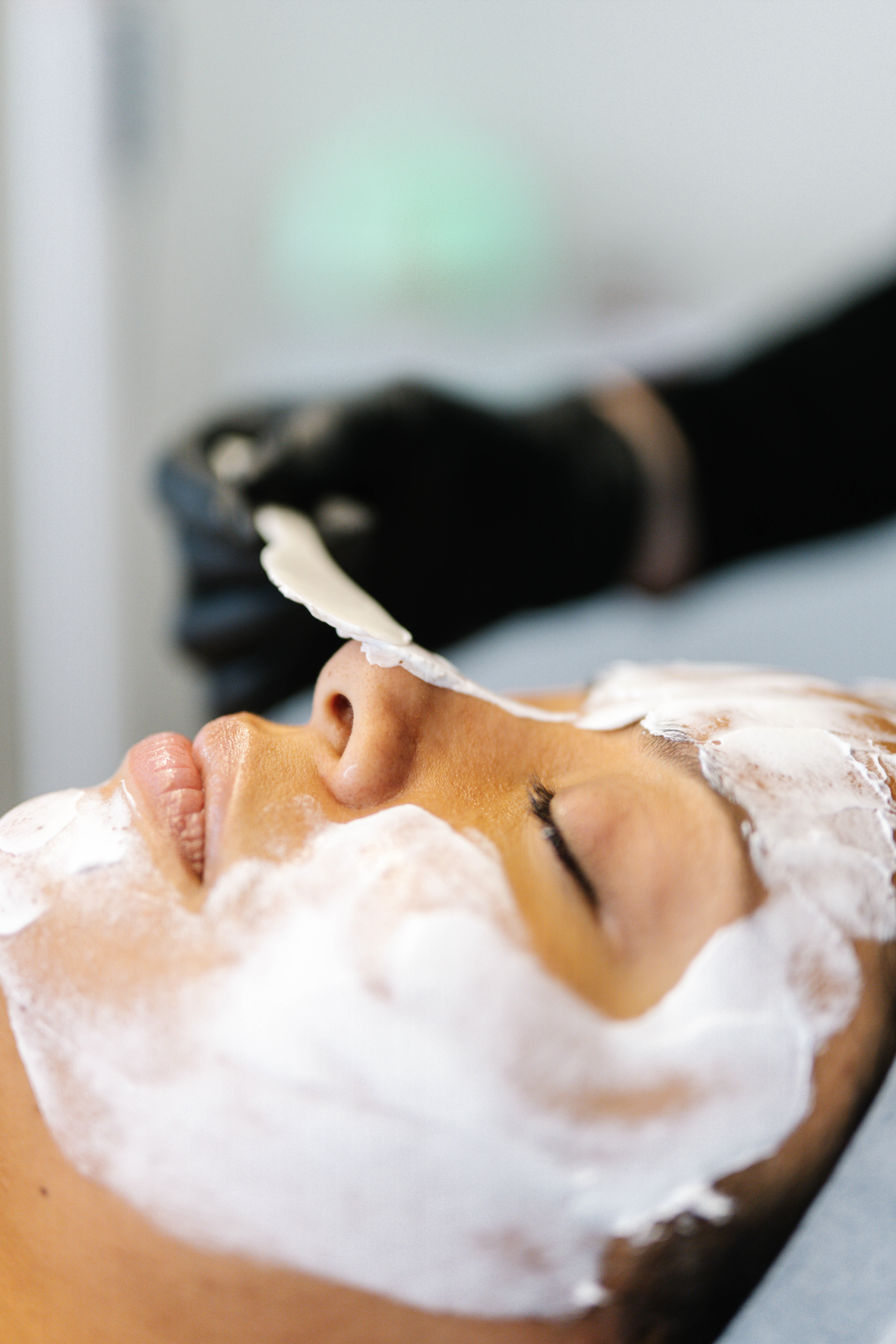 A woman lies on her back with her eyes closed, receiving a facial treatment with a foamy white substance, while a person wearing black gloves applies the treatment.