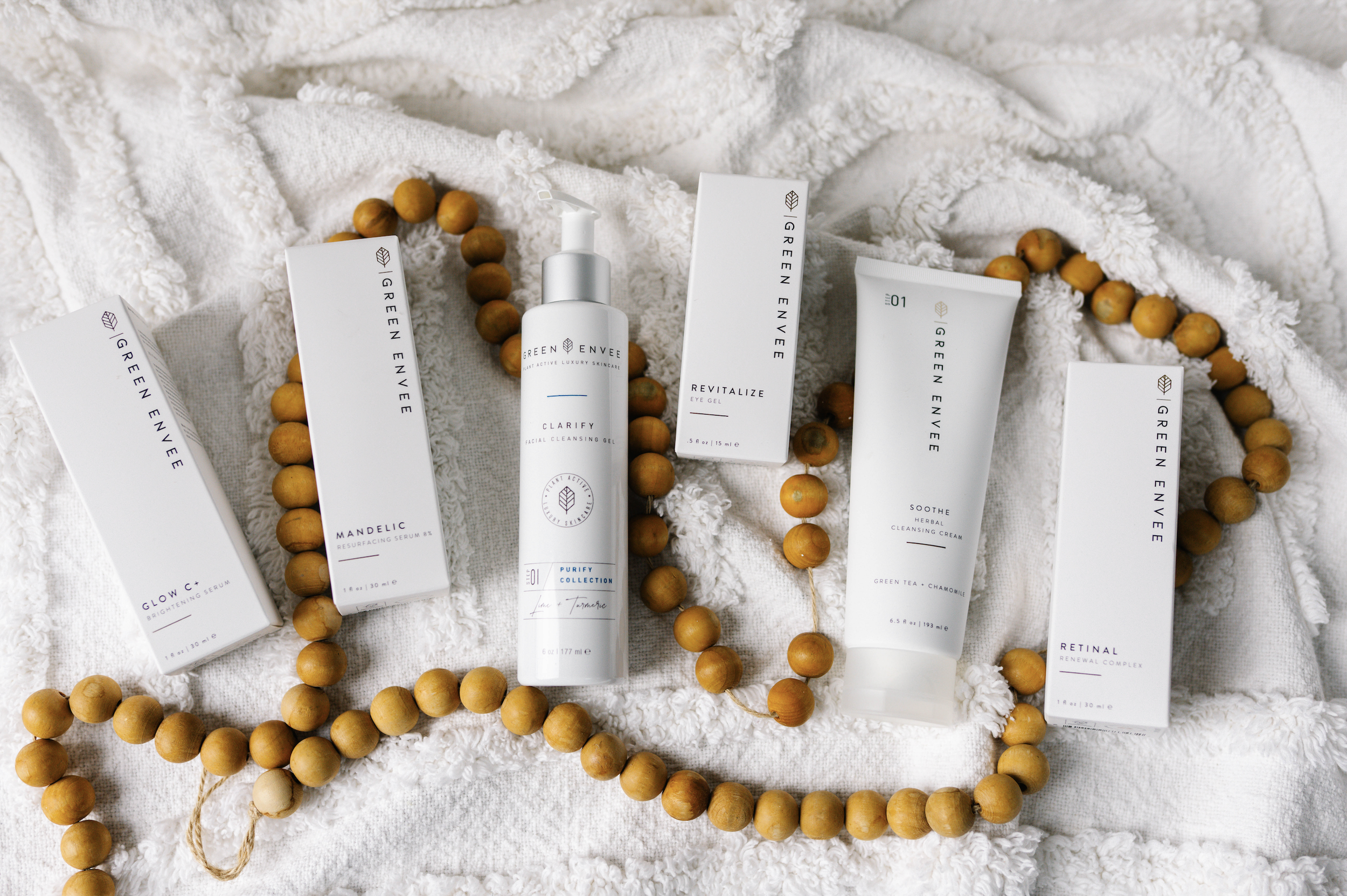 Skincare products from Green Envee including Purify Facial Cleansing Gel, Revitalize Eye Gel, Soothe Herbal Cleansing Cream, and Resurfacing Serum, arranged on a white textured towel with wooden beads.