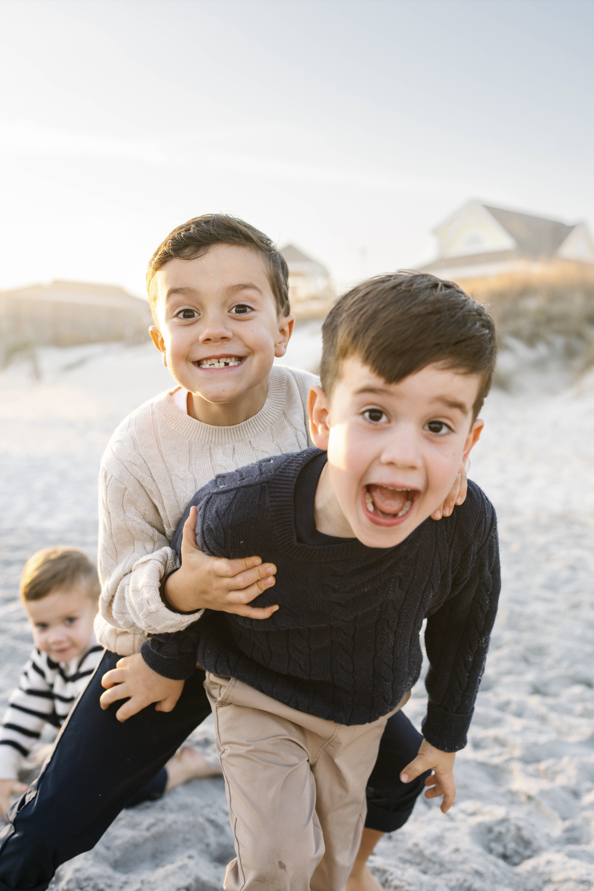 Three smiling children playing on a sandy beach, with two boys in the foreground and a girl in the background. The boys are stacked, with the older boy on his hands and knees and the younger boy on top, leaning forward. The girl is sitting on the san
