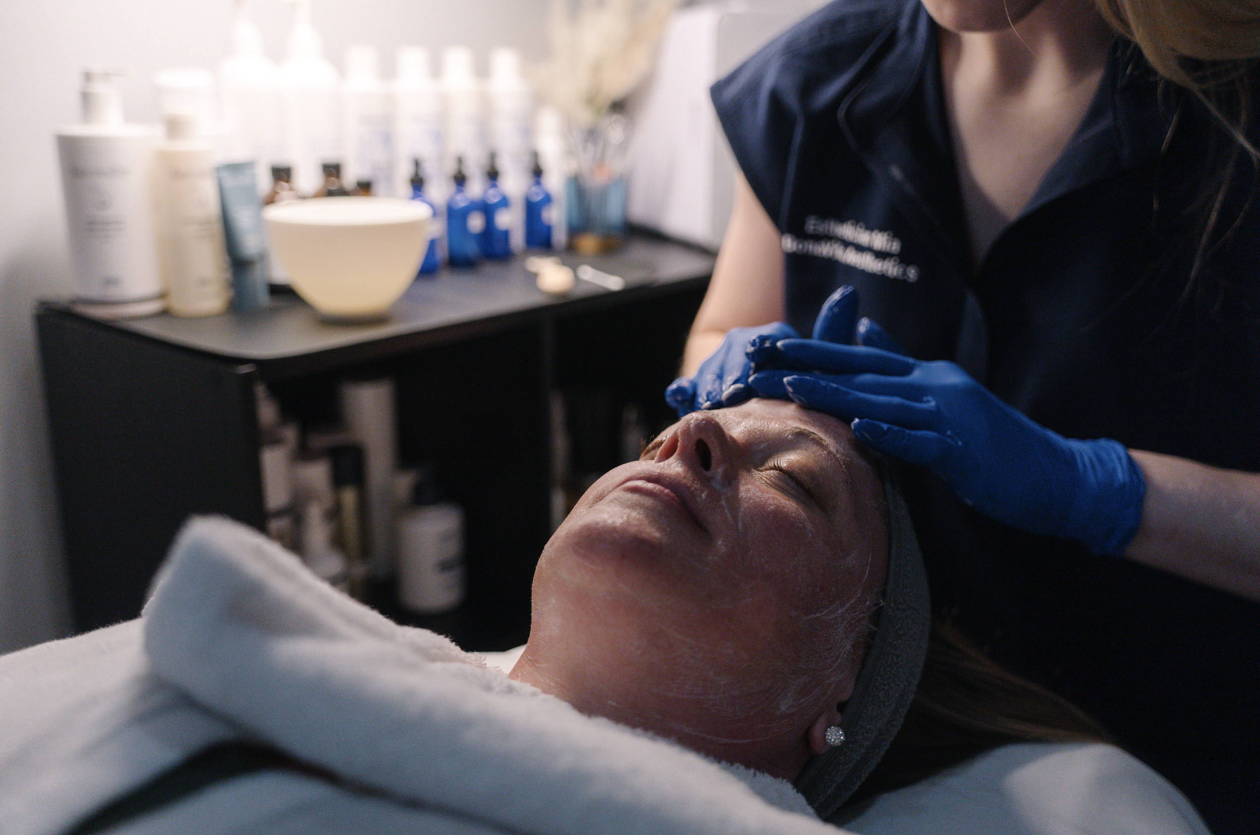 A woman receiving a facial treatment at a spa from an esthetician in a dark uniform with blue gloves, with skincare products on a shelf behind them.